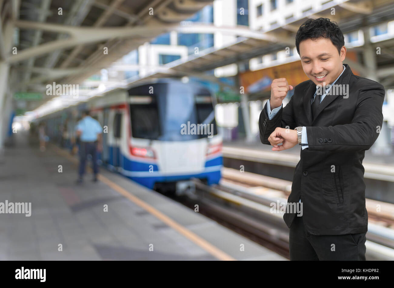 Young businessman looking at watch which cheerful action on abstract Blurred photo of sky train at station with security guard, business rush hour con Stock Photo