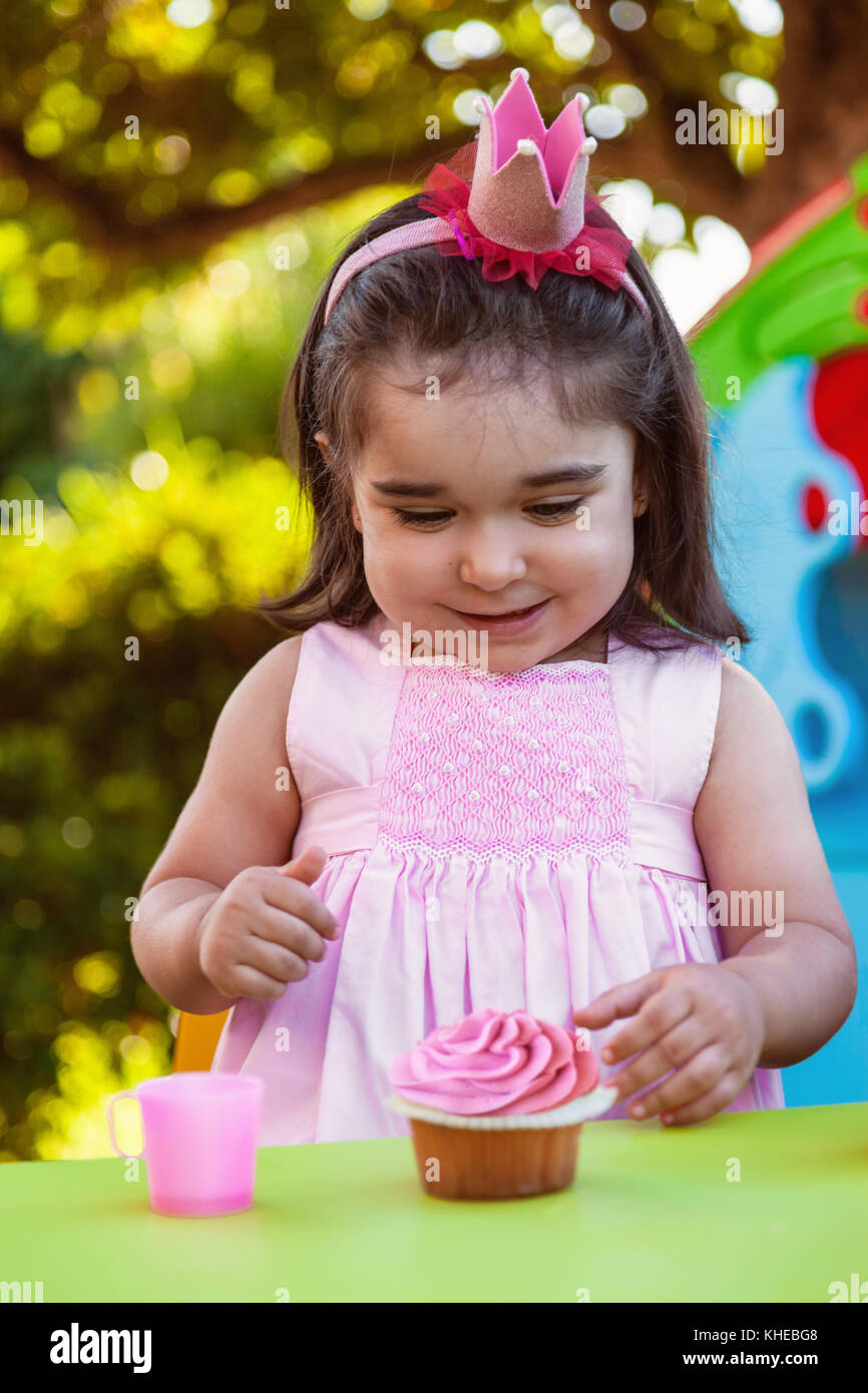Baby toddler girl in outdoor party at garden, happy and smiling at cupcake with sweet tooth expression. Pink dress and queen or princess crown. Stock Photo