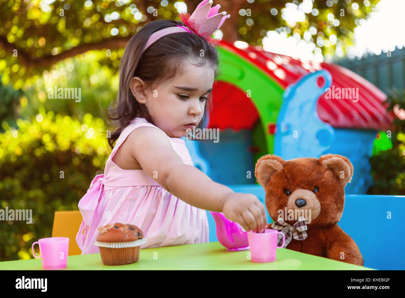 Baby toddler girl playing in outdoor tea party serving her best friend Teddy Bear with candy gummy. Pink dress and queen or princess crown. Stock Photo