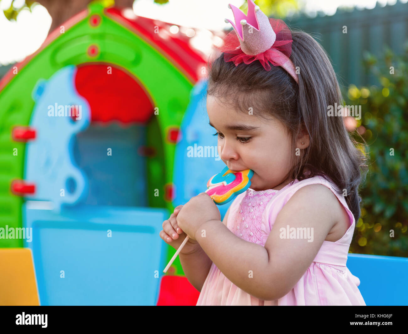 Baby toddler girl eating a large colorful lollipop dressed in pink dress as princess or queen with crown, playing outdoor in garden with playhouse Stock Photo