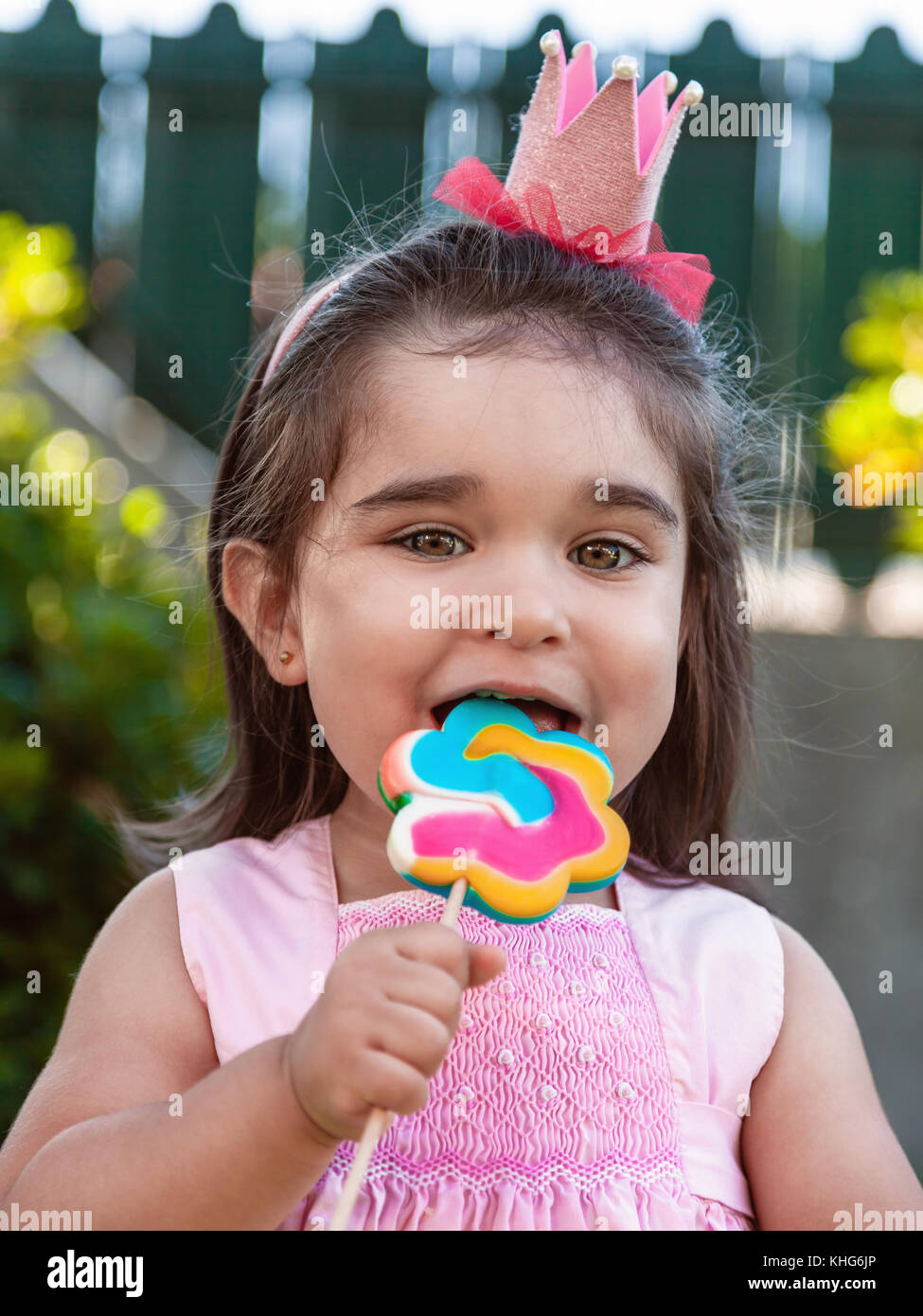 Happy baby toddler girl eating and biting a large colorful lollipop dressed in pink dress as princess or queen with crown, playing outdoor in garden Stock Photo