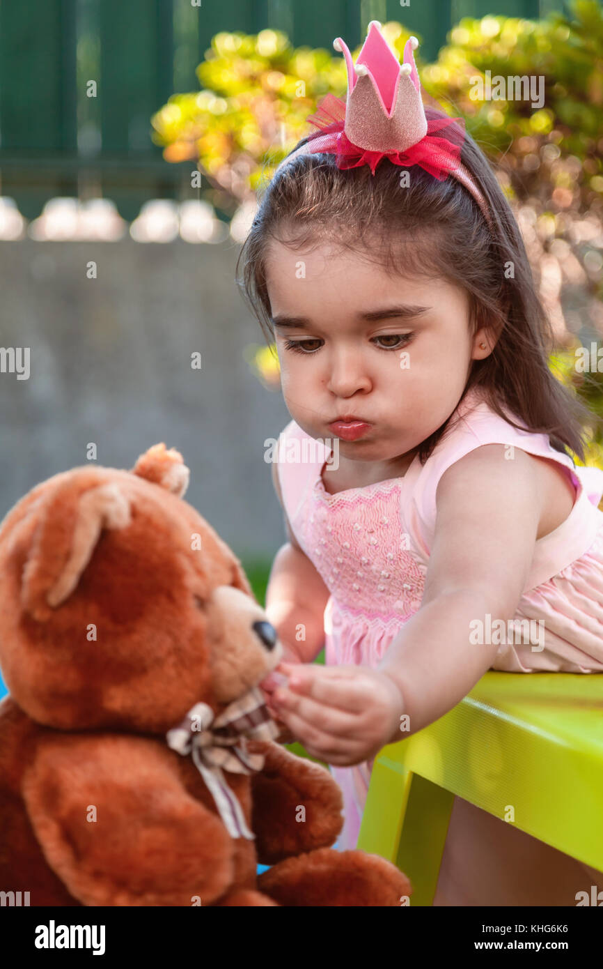 Baby toddler girl, mouth full of candies playing in a tea party feeds best friend Teddy Bear with candy gummy. Pink dress and queen or princess crown Stock Photo
