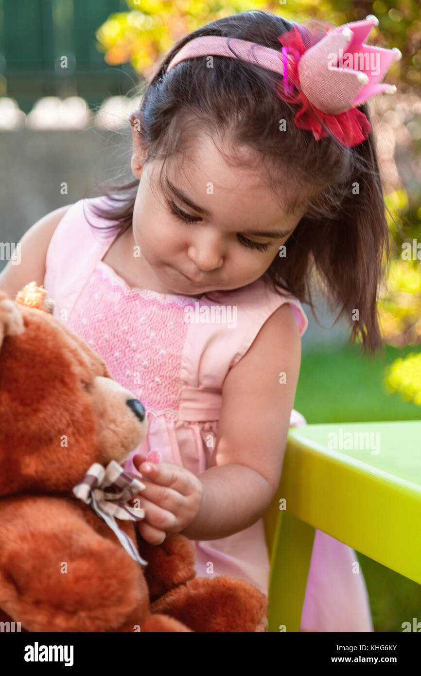Baby toddler girl, playing in a tea party feeds best friend bff Teddy Bear with candy gummy. Pink dress and queen or princess crown Stock Photo