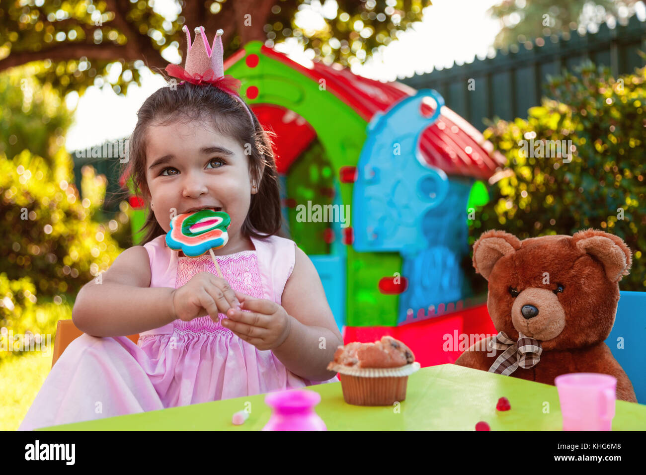 Baby toddler girl playing in outdoor tea party eating and biting a large lollipop with best friend Teddy Bear. Pink dress and queen or princess crown. Stock Photo