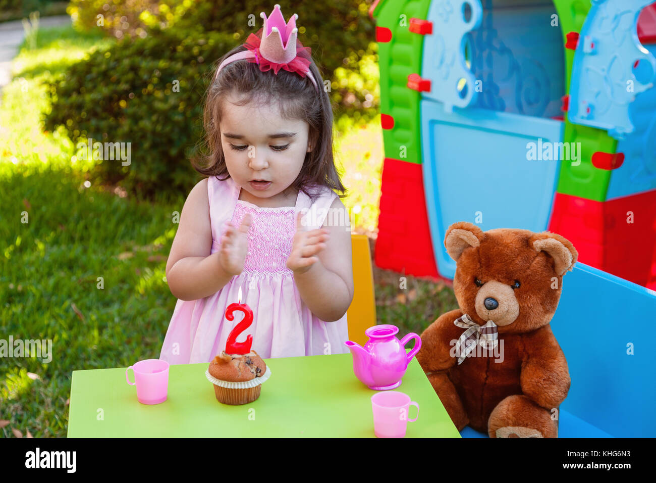 Baby toddler girl in outdoor second birthday party clapping hands at cake with Teddy Bear as best friend, playhouse and tea set. Pink dress and crown. Stock Photo