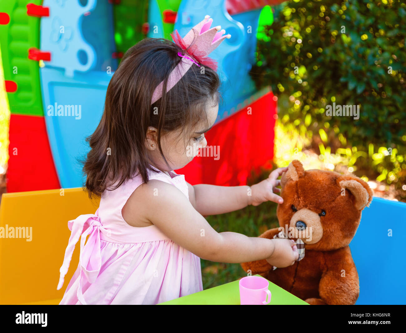 Baby toddler girl playing in outdoor tea party feeding her best friend Teddy Bear with candy gummy. Pink dress and queen or princess crown. Stock Photo