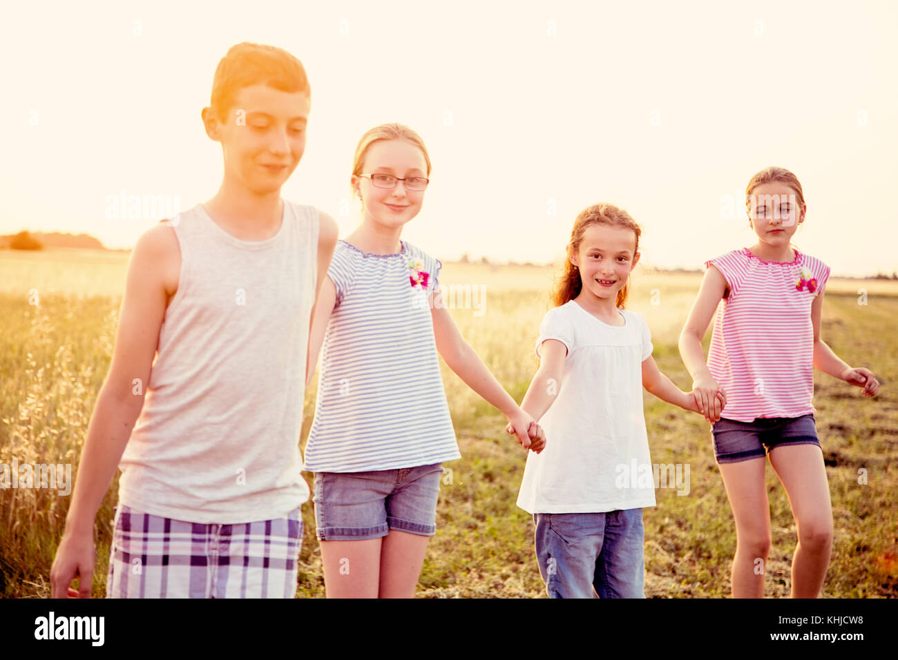 Four young caucasian kids holding hands, walking at cultivated countryside field, at summer sunset, wearing casual summer clothes Stock Photo