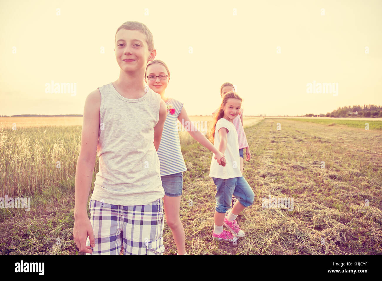 Four young caucasian kids holding hands, walking at cultivated countryside field, at summer sunset, wearing casual summer clothes Stock Photo