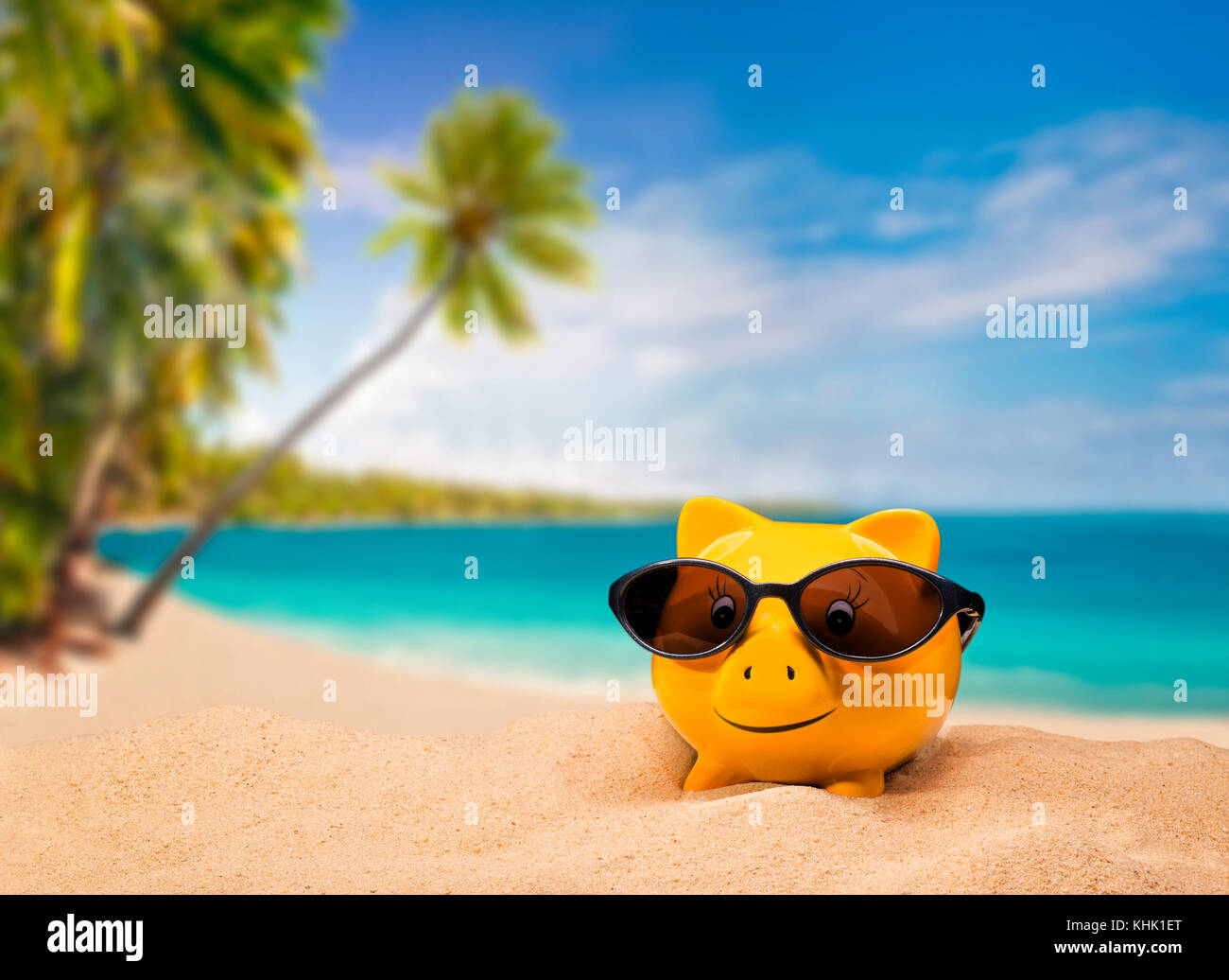 Yellow piggy bank with sunglasses with a tropical beach in the background Stock Photo