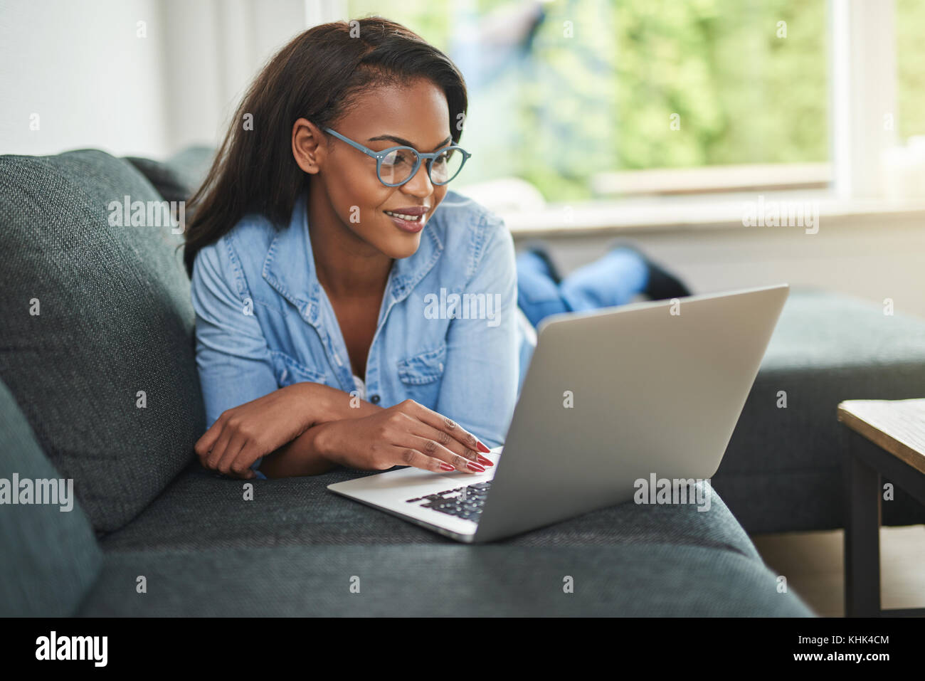 Smiling young African woman lying down on her living room sofa using a ...