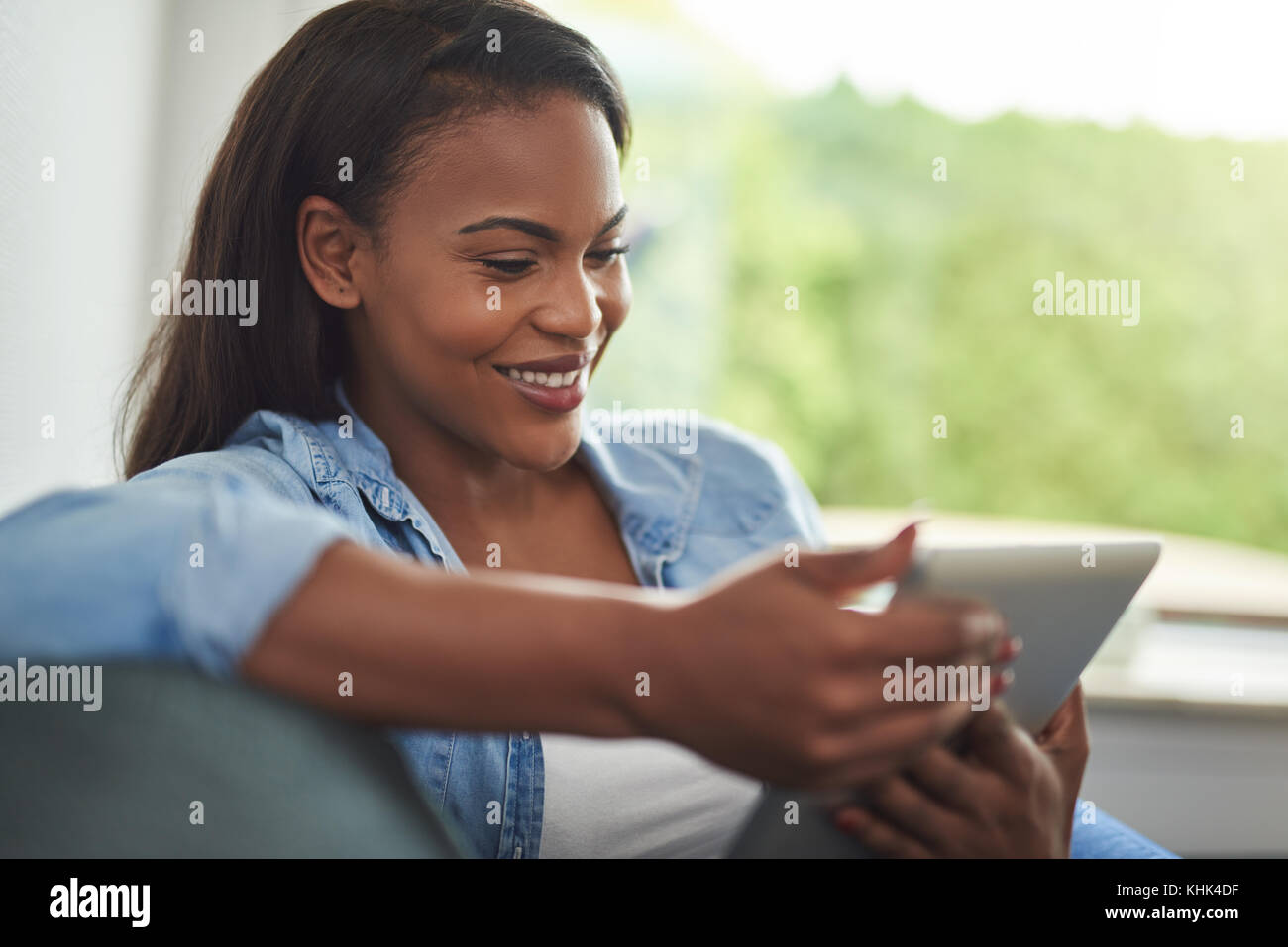 Smiling young African woman relaxing on her living room sofa browsing ...