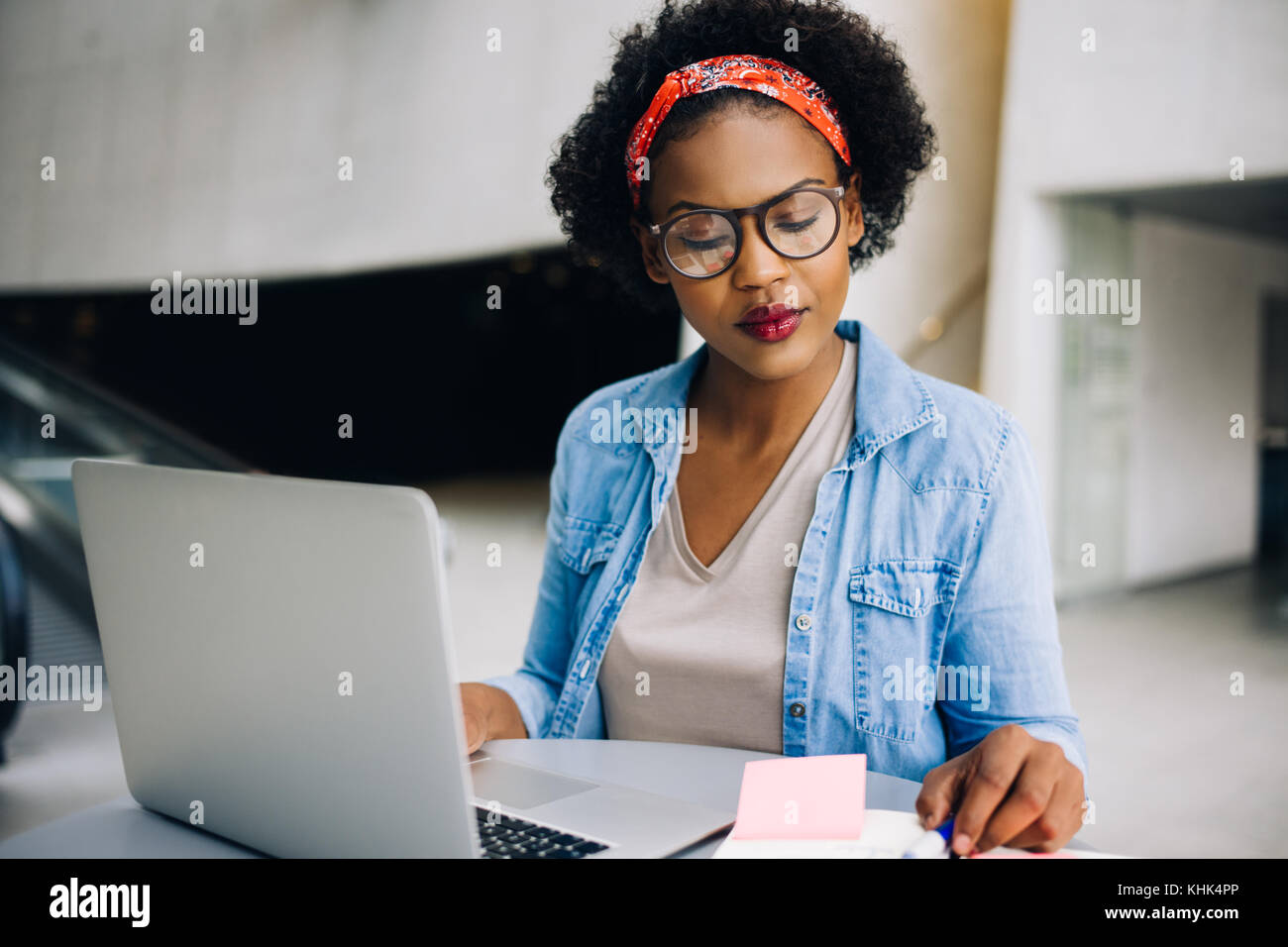 Confident young African female entrepreneur sitting at a table in a modern office building lobby working on a laptop and reading notes in her planner Stock Photo