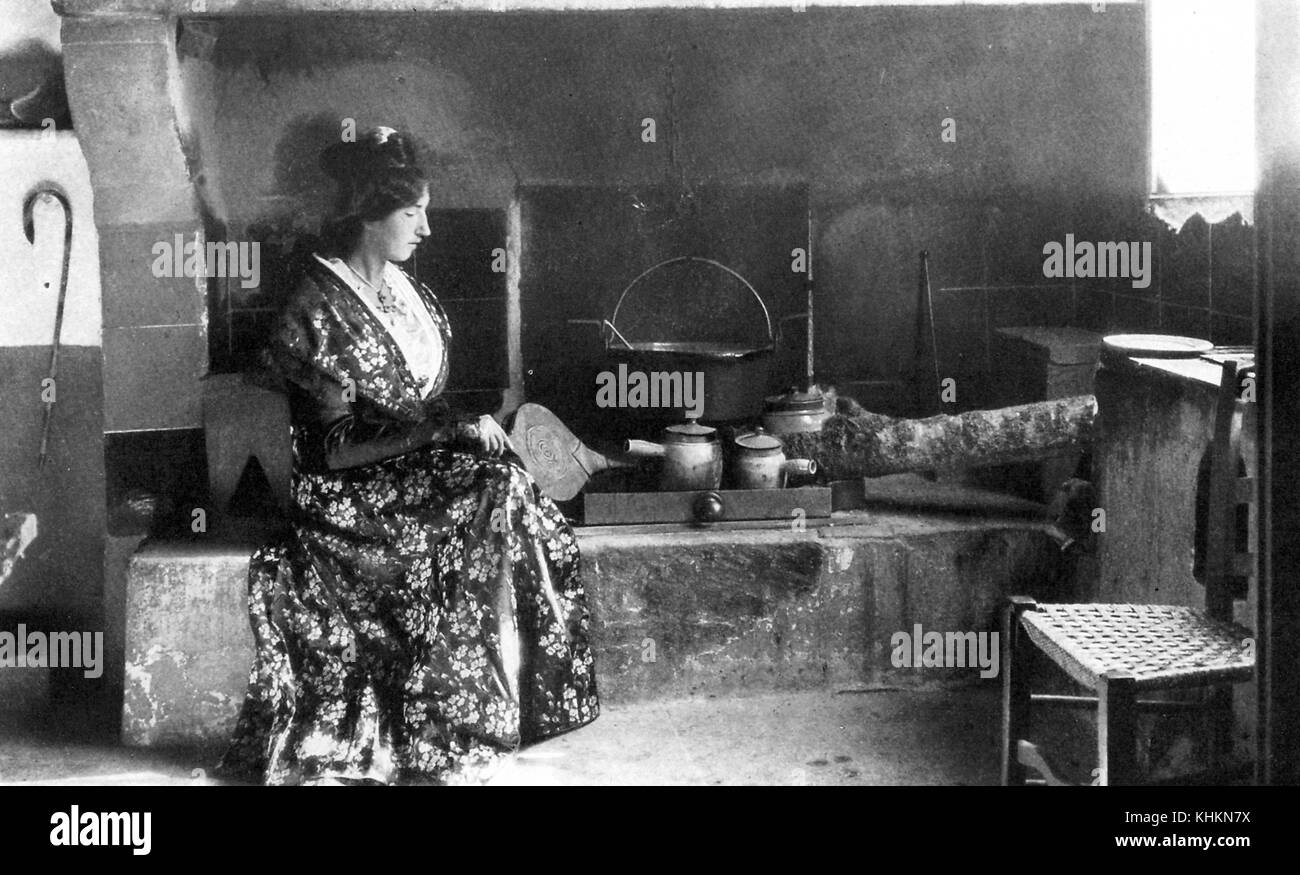 A photograph of a woman posed while wearing traditional clothing, she was shown cooking in a farmhouse kitchen and using hand bellows to provide air for a fire in a large open fireplace, a kitchen like this was in every farmhouse and used to provide food for the farmer, his family, and laborers, Camargue, France, July, 1922. Stock Photo
