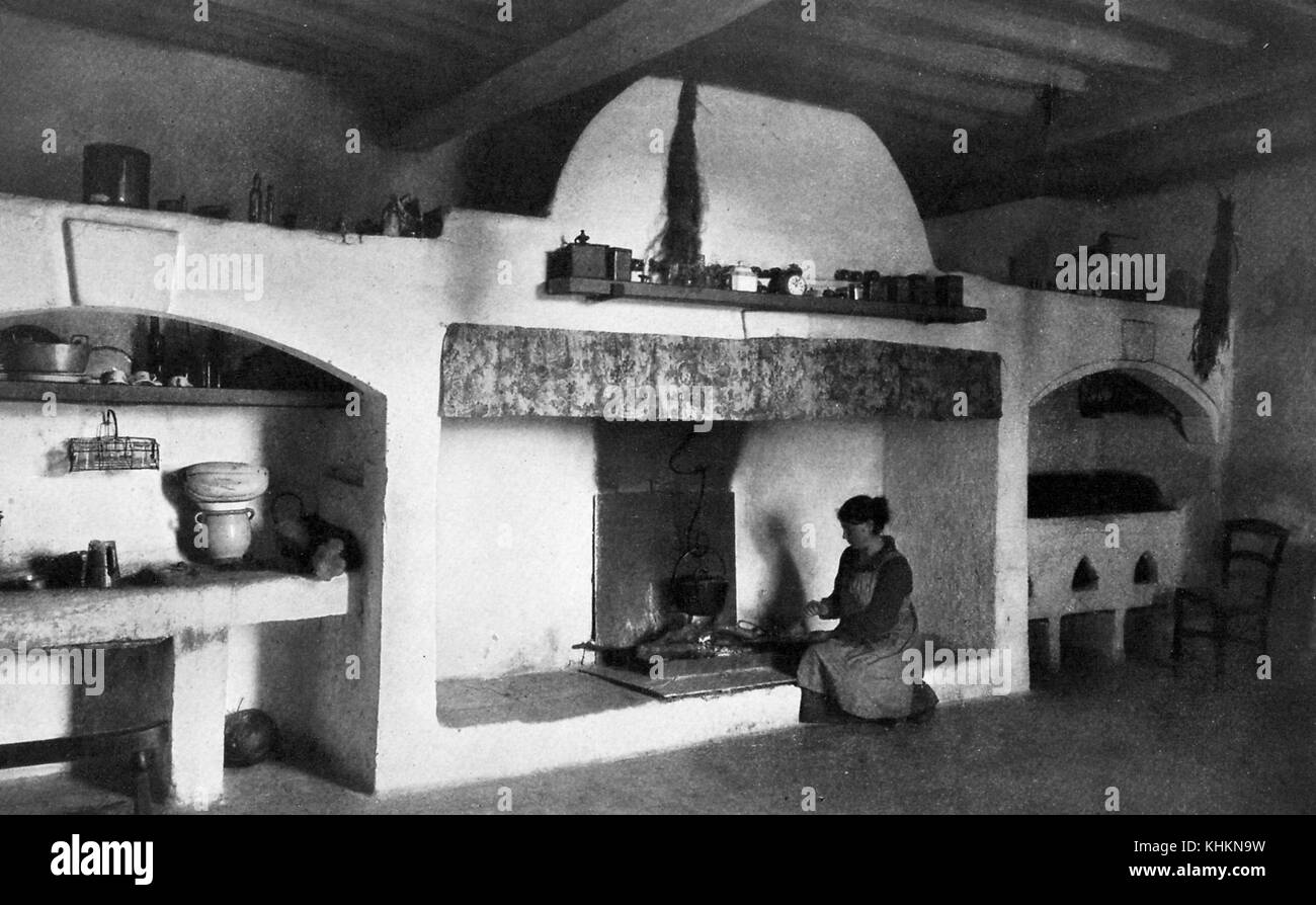 A photograph that shows a housewife cooking in the home of a herder, the large open fireplace where she is knelt down cooking is located centrally in the room, on the mantle are a number of kitchen items, to the left of the fireplace is an area that includes the washing basin and cooking utensils, Camargue, France, July, 1922. Stock Photo