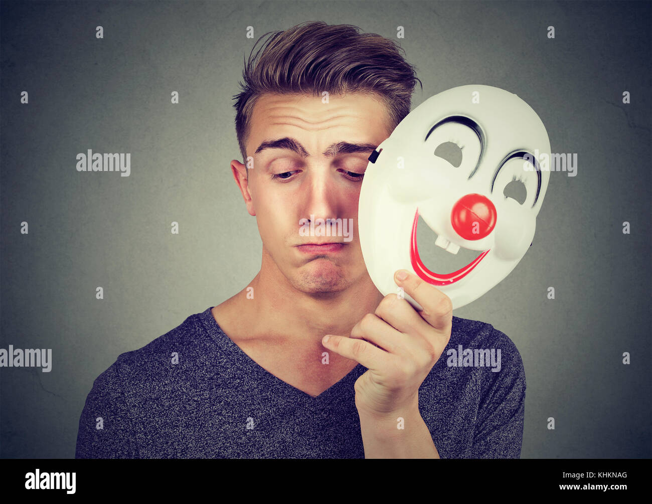 Young sad man taking off happy clown mask isolated on gray background. Human emotions. Stock Photo