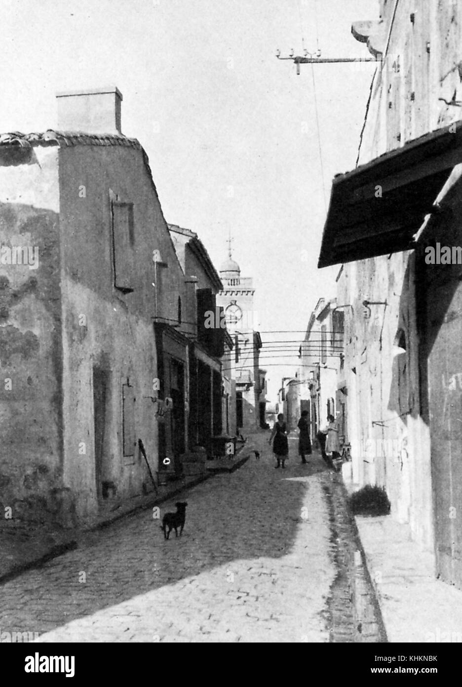 A photograph depicting a typical street in Les Saintes Maries de la Mer, which is the capital city of the region, the narrow granite paved street is lined by short, modest stone houses that are densely packed, roofs made of tiles can be seen on tops of the houses, women are shown walking and talking in the street, a stray dog stands watching them, Les Saintes Maries de la Mer, Camargue, France, July, 1922. Stock Photo
