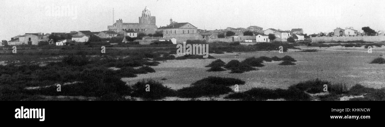 A photograph of a landscape that shows a large, fortified cathedral towering over the other buildings in the town, the foreground is made up of sand and short beach grass, the cathedral once served as protection from pirates, Saintes Maries de la Mer, Camargue, France, July, 1922. Stock Photo