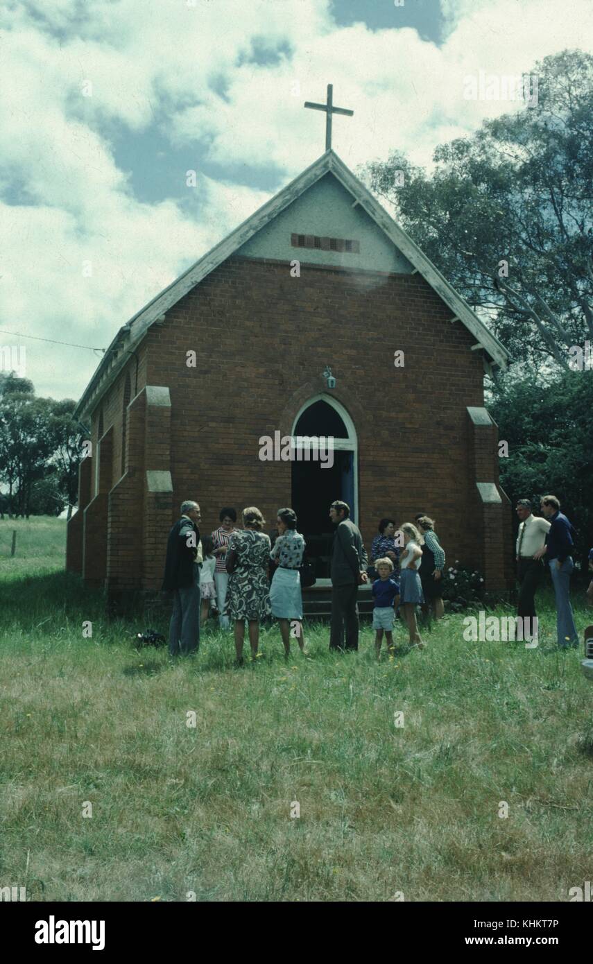 A photograph of a group of families gathered outside of the entrance to a church, the women and children are wearing summer dresses and shorts while the men wear long pants, button down shirts and some wear suits, the church is small and topped with a cross, the lawn surrounding the church is unkempt, 1965. Stock Photo