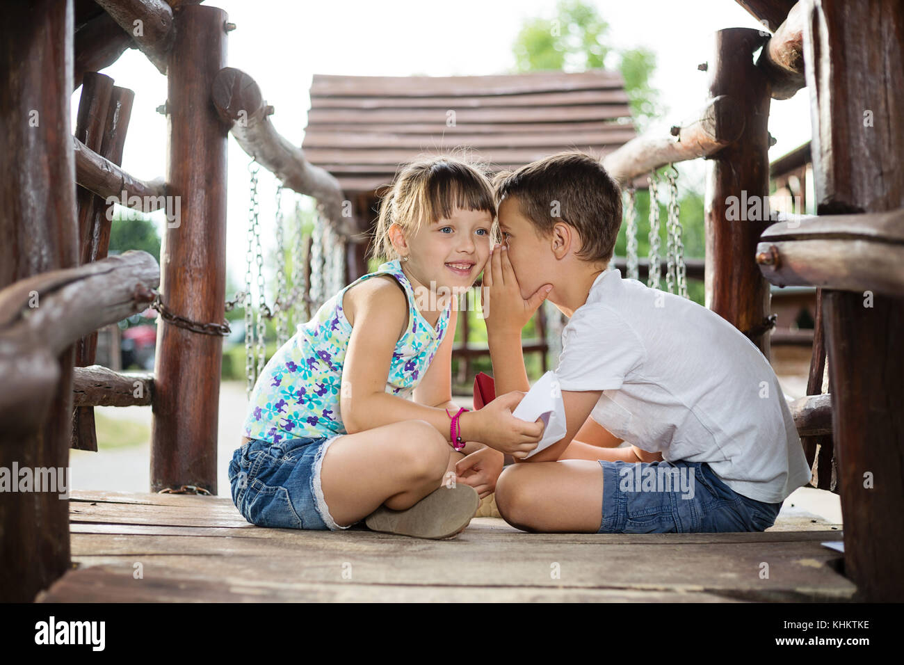 Happy two caucasian siblings sittign in a wooden house outdoors on summer day,  holding a paper boats in their hands, boy whispering a secret into gir Stock Photo
