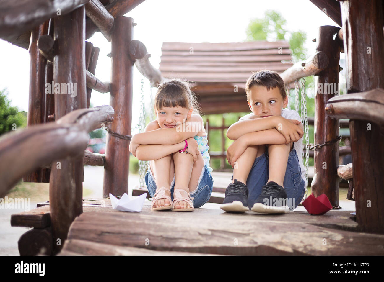 Two young caucasian children sitting next to each other with bent knees in a wooden house, outdoors on sunny summer day. They look innocent Stock Photo