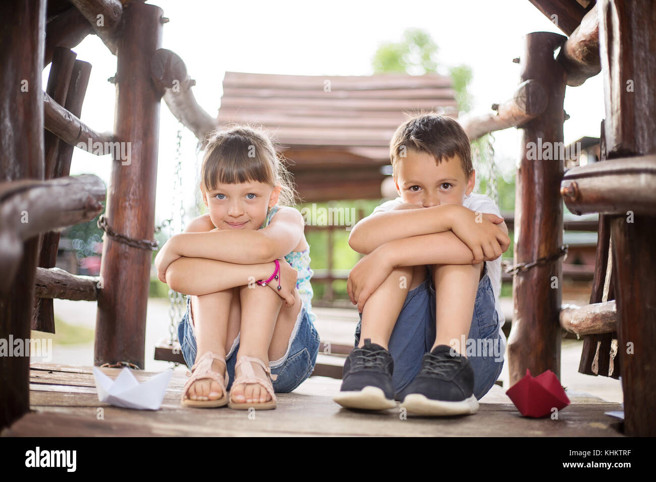 Two young caucasian children sitting next to each other with bent knees in a wooden house, outdoors on sunny summer day. They look innocent Stock Photo