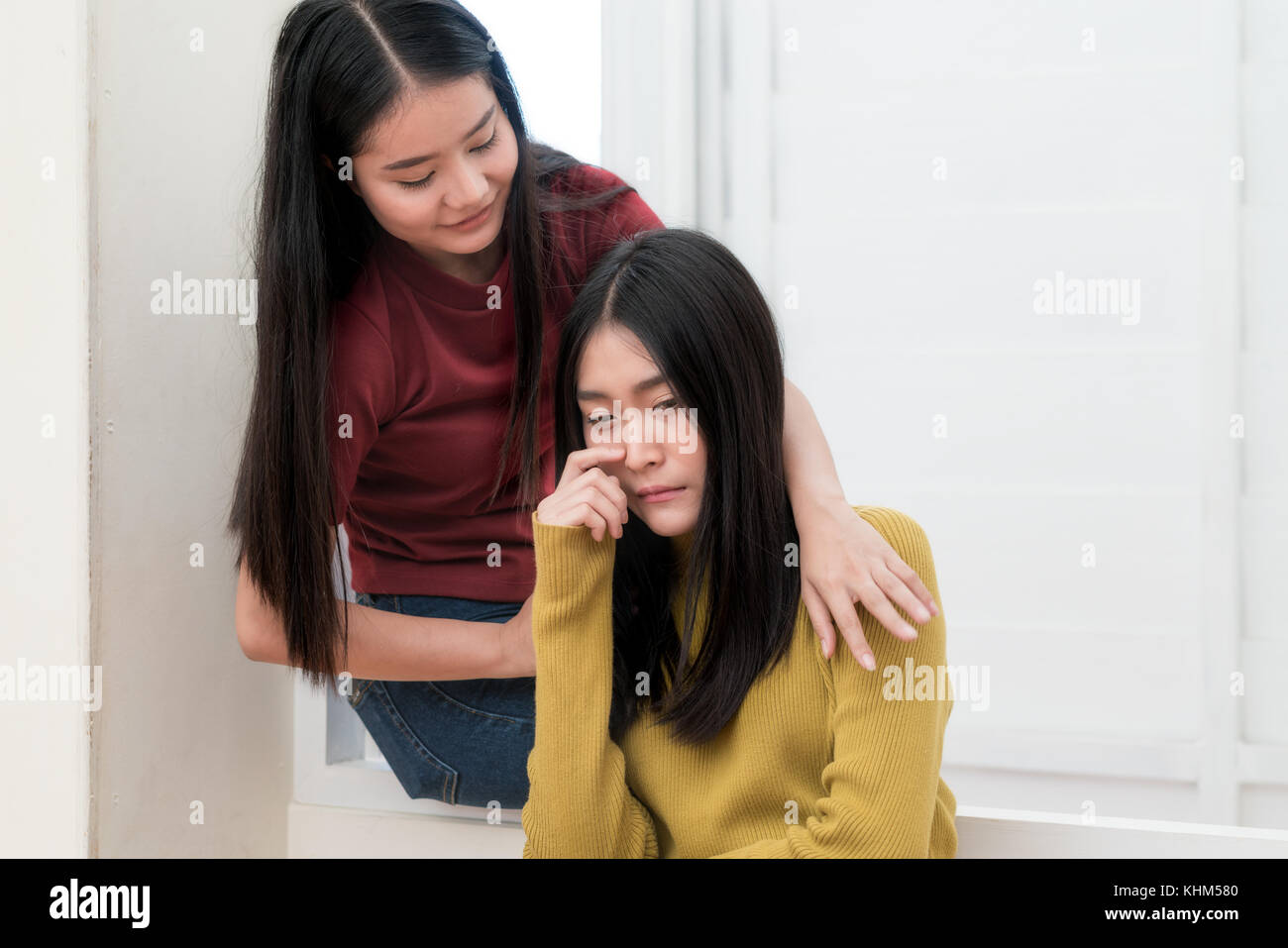 Beautiful Asian woman hugging depressed friend while sitting on window at home Stock Photo