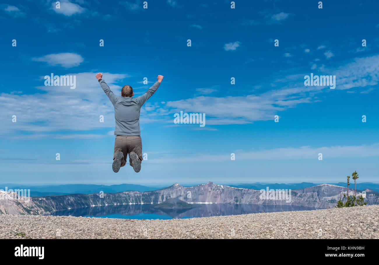 Man Jumps on Rocky Overlook to Crater Lake on Summer morning Stock Photo