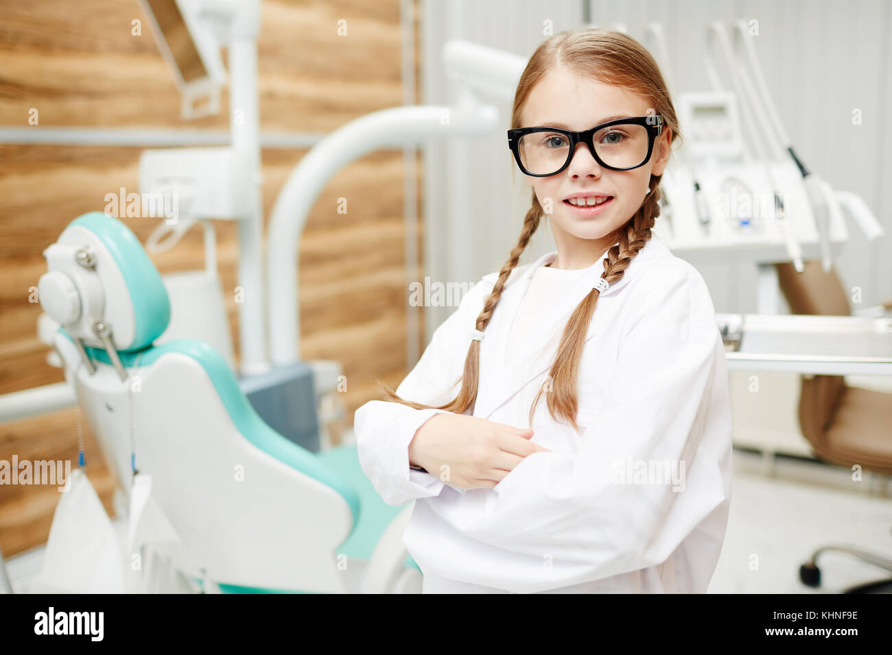 Cute child in eyeglasses and whitecoat crossing her arms on chest in dental office Stock Photo
