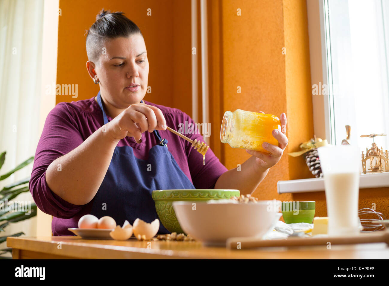 Close up photo of young female holding a jar of organic honey above a green bowl with one hand, getting spoon of honey from the jar Stock Photo