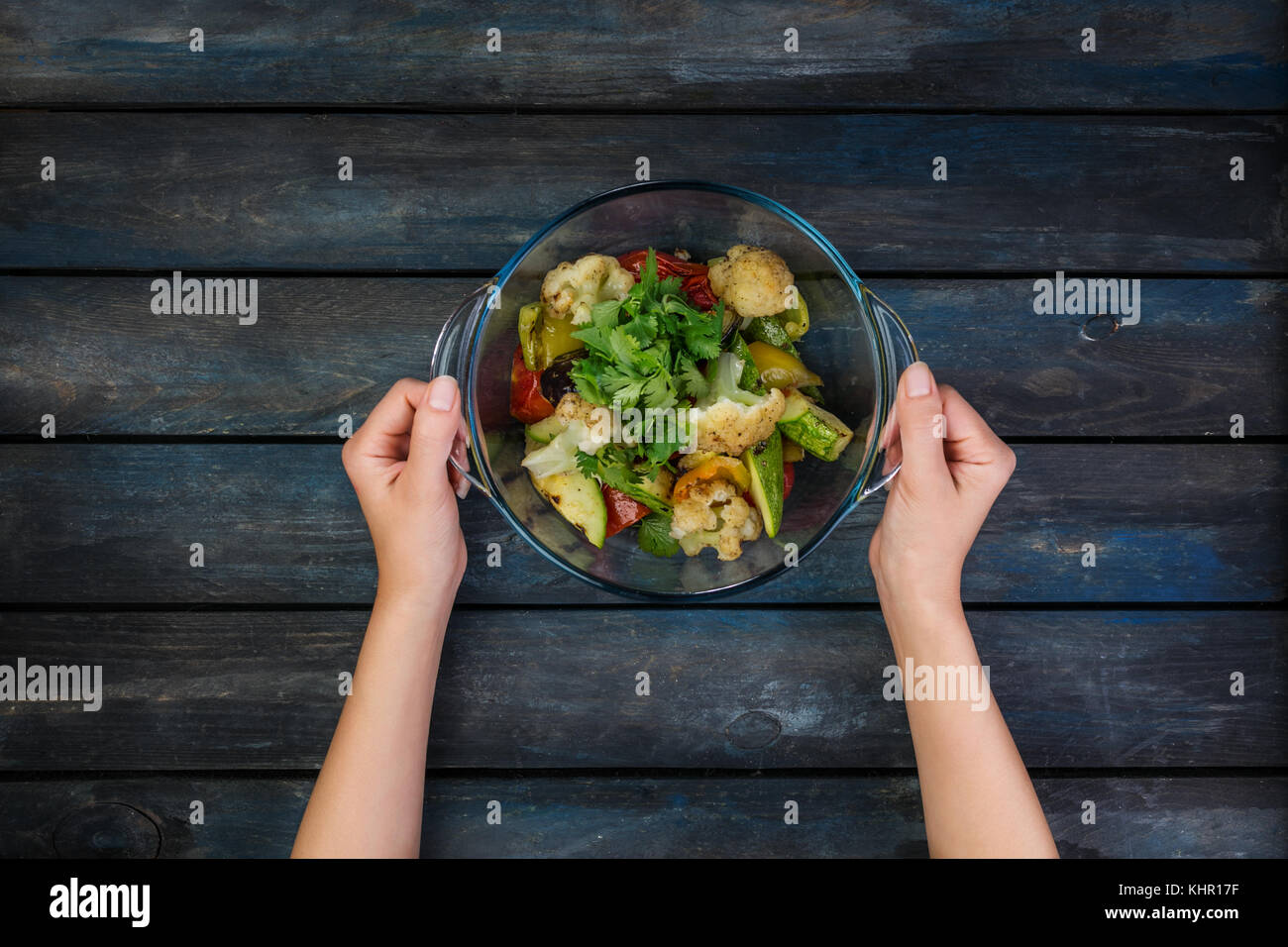 Female hands holding the plate with grilled vegetables salad with zucchini, eggplant, onions, peppers and tomato. On a colored wooden background. Top  Stock Photo