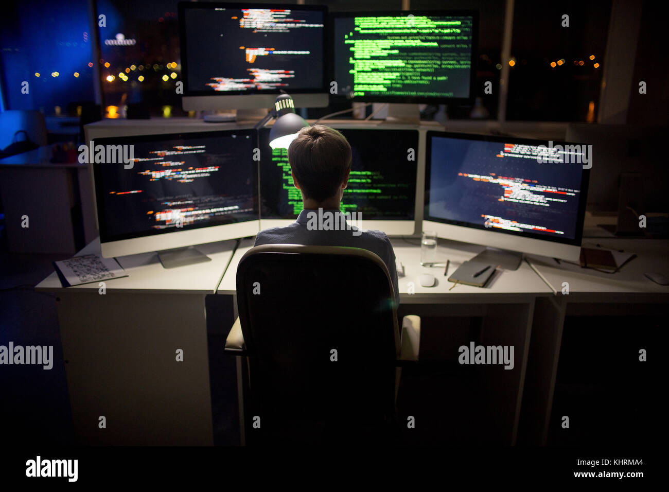 Rear view of hard-working programmer with stylish haircut sitting in front of computer and writing code, interior of dim open plan office on backgroun Stock Photo