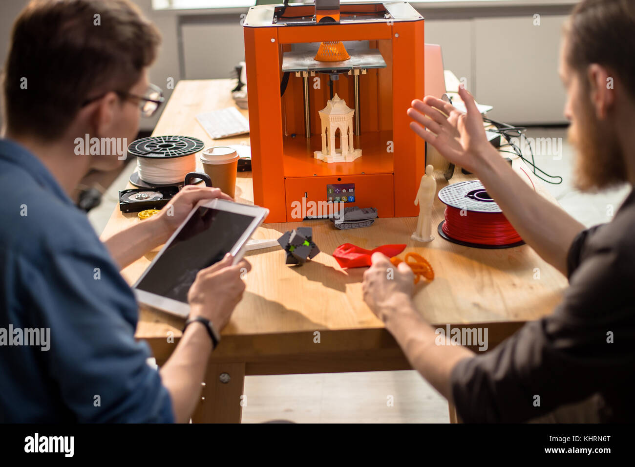 Back view of talented young engineers sitting at desk and carrying out quality inspection of prototype made with help of 3D printer, interior of moder Stock Photo