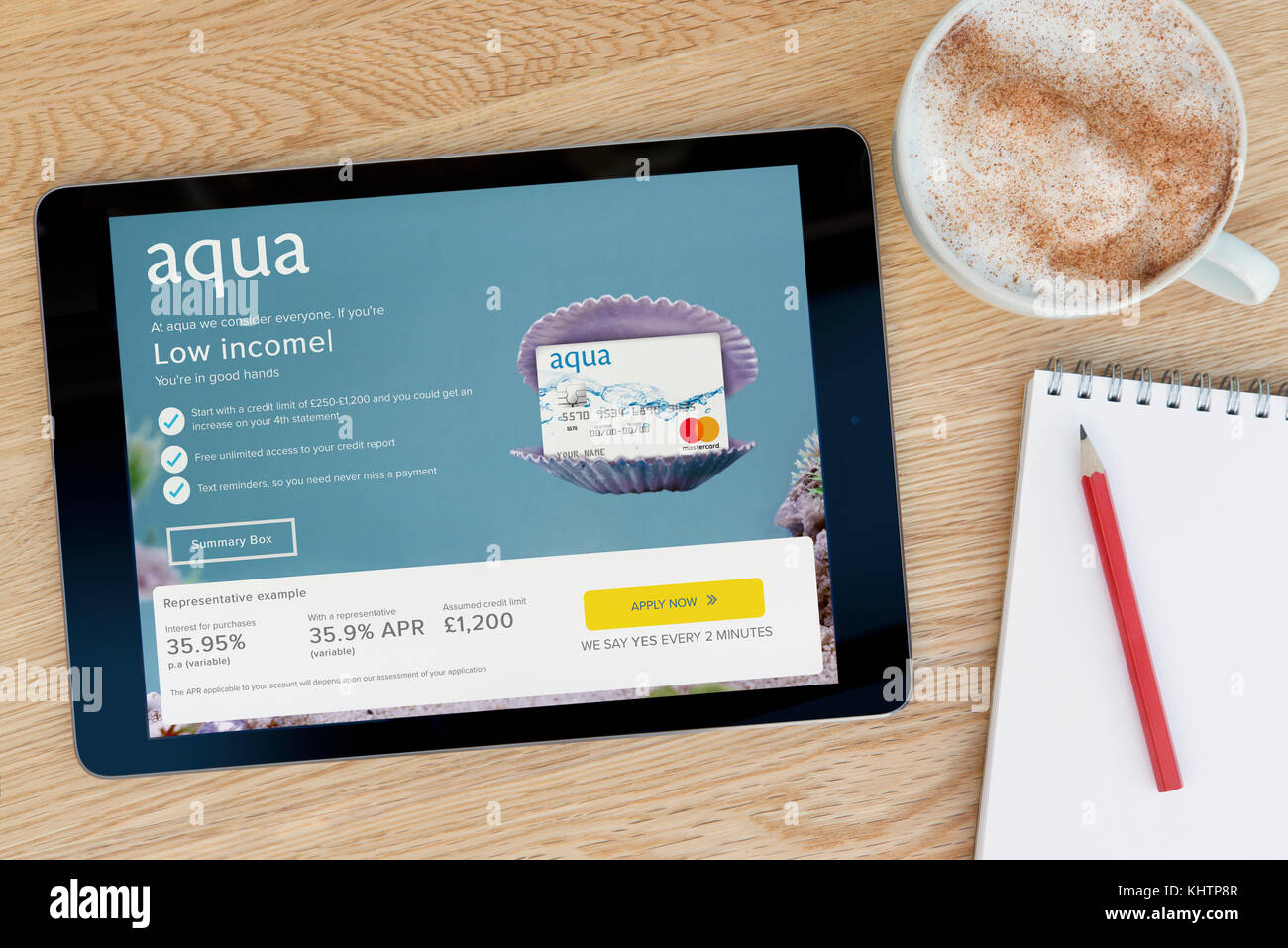 The Aqua Card website features on an iPad tablet device which rests on a wooden table beside a notepad and pencil and a cup of coffee (Editorial only) Stock Photo