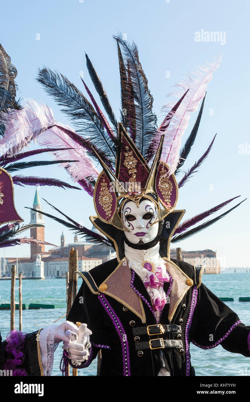 Venice Carnival 2017, man in elegant feathered headdress and mask posing in front of the lagoonand San Giorgio Maggiore Stock Photo