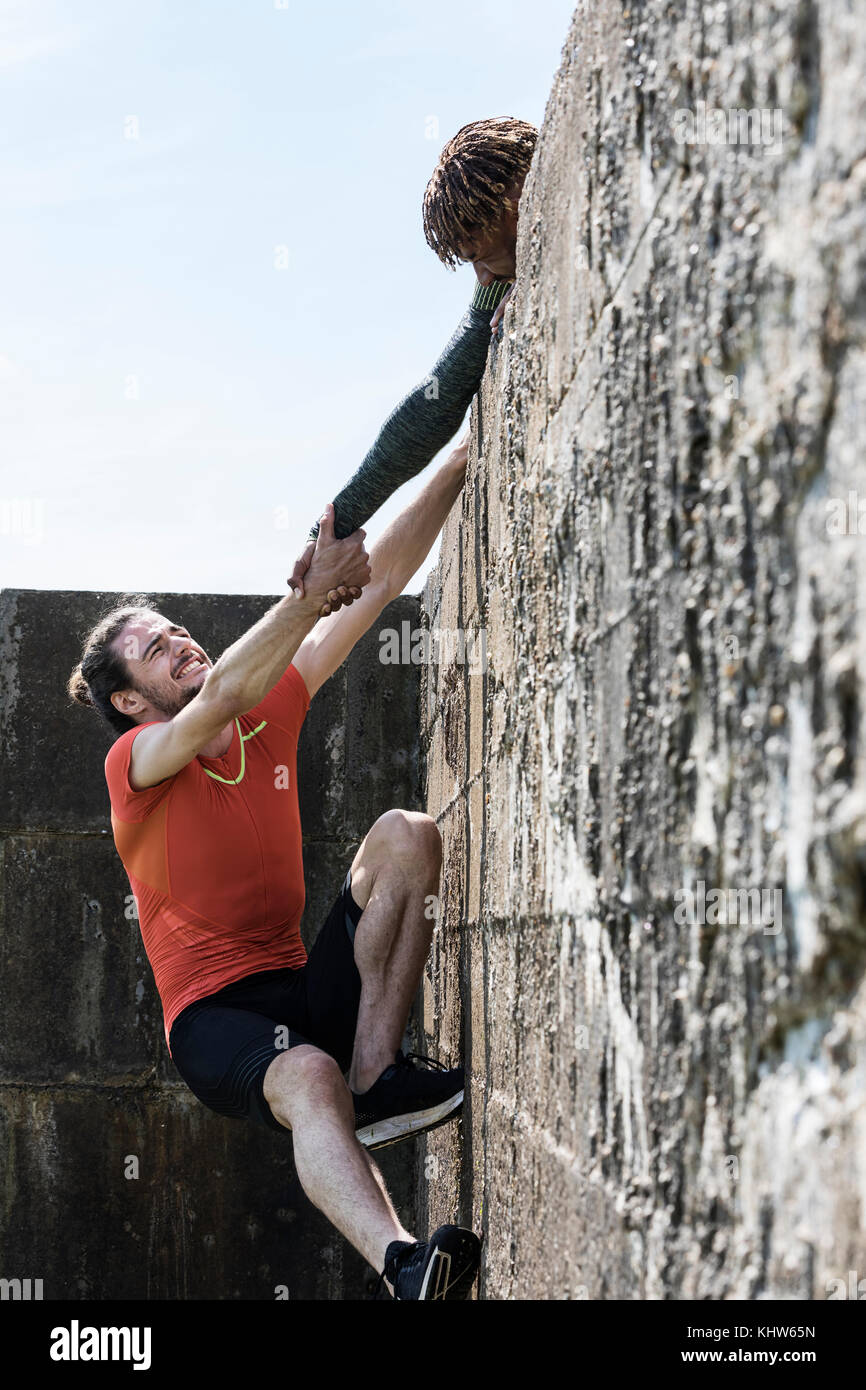 Young male free climber at top of sea wall helping friend climb up Stock Photo