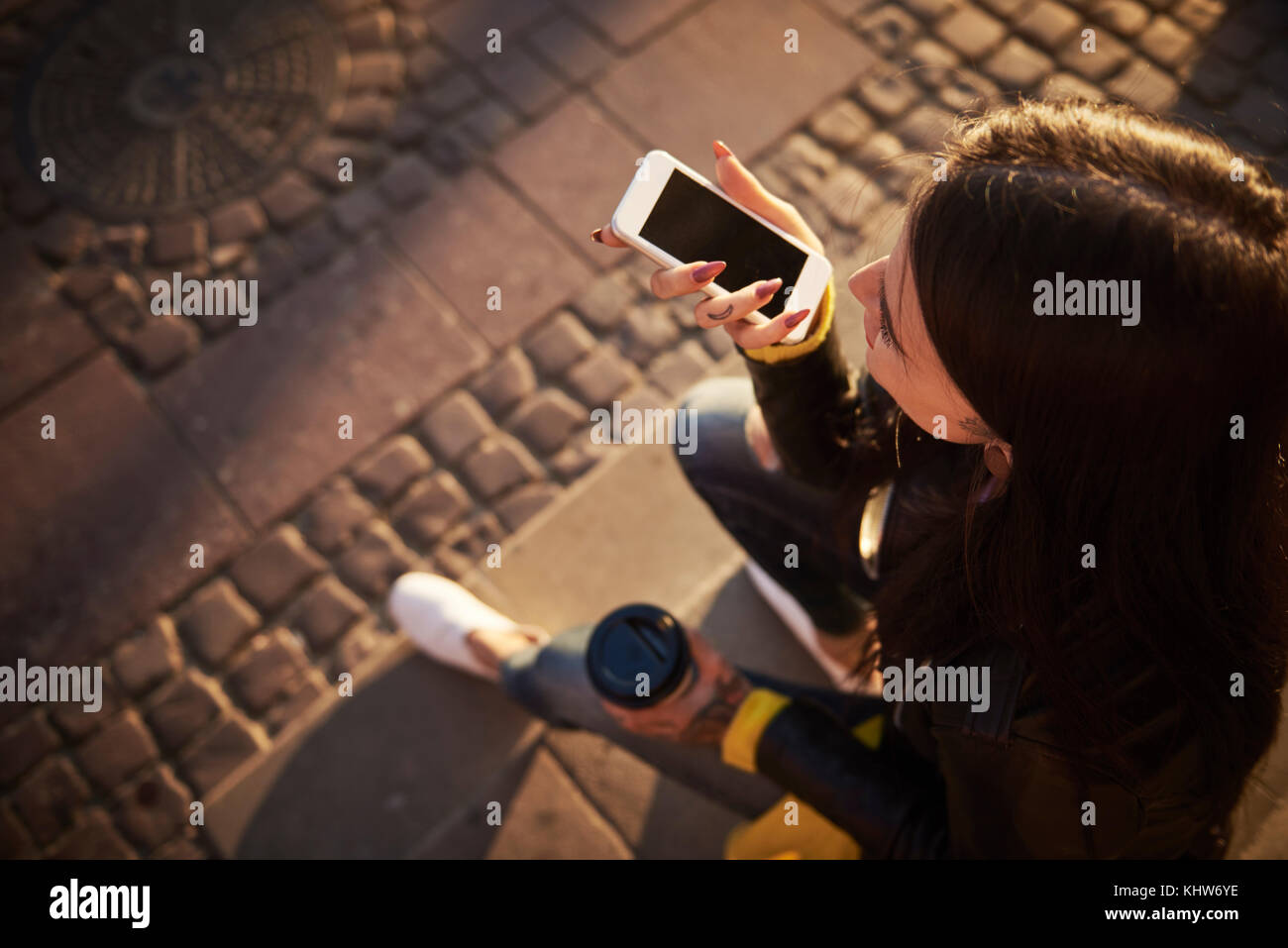 Young woman sitting outdoors, holding coffee cup, using smartphone, tattoos on hands, elevated view Stock Photo