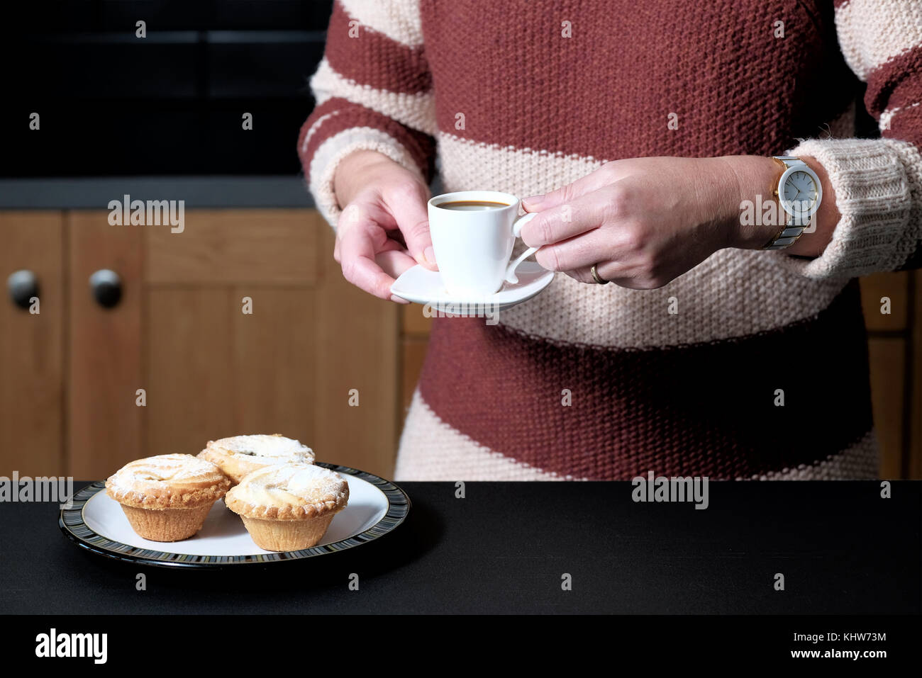 A smart but casually dressed woman holding an espresso coffee whilst standing in her kitchen at home with a plate of mince pies Stock Photo
