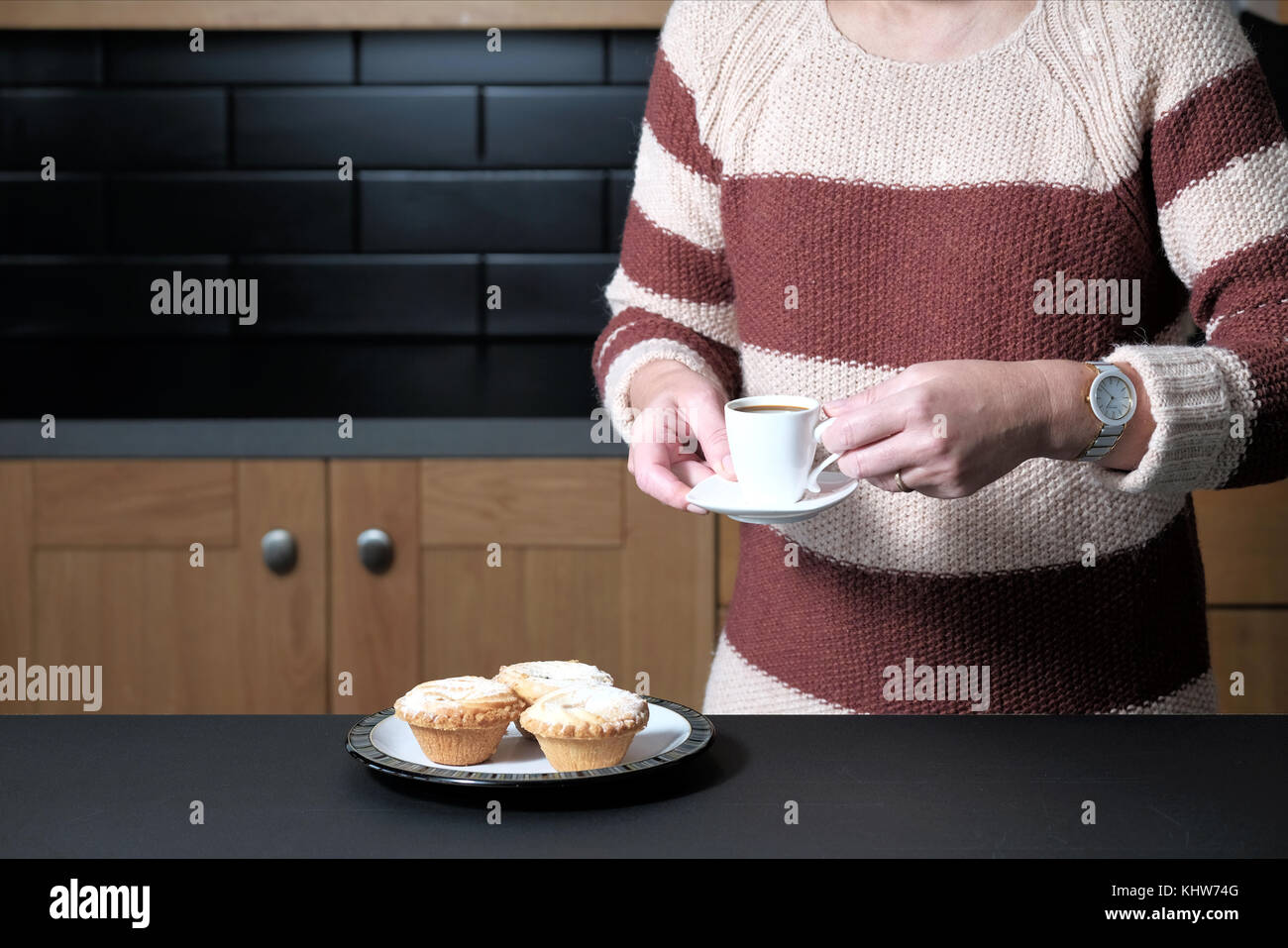 A smart but casually dressed woman holding an espresso coffee whilst standing in her kitchen at home with a plate of mince pies Stock Photo