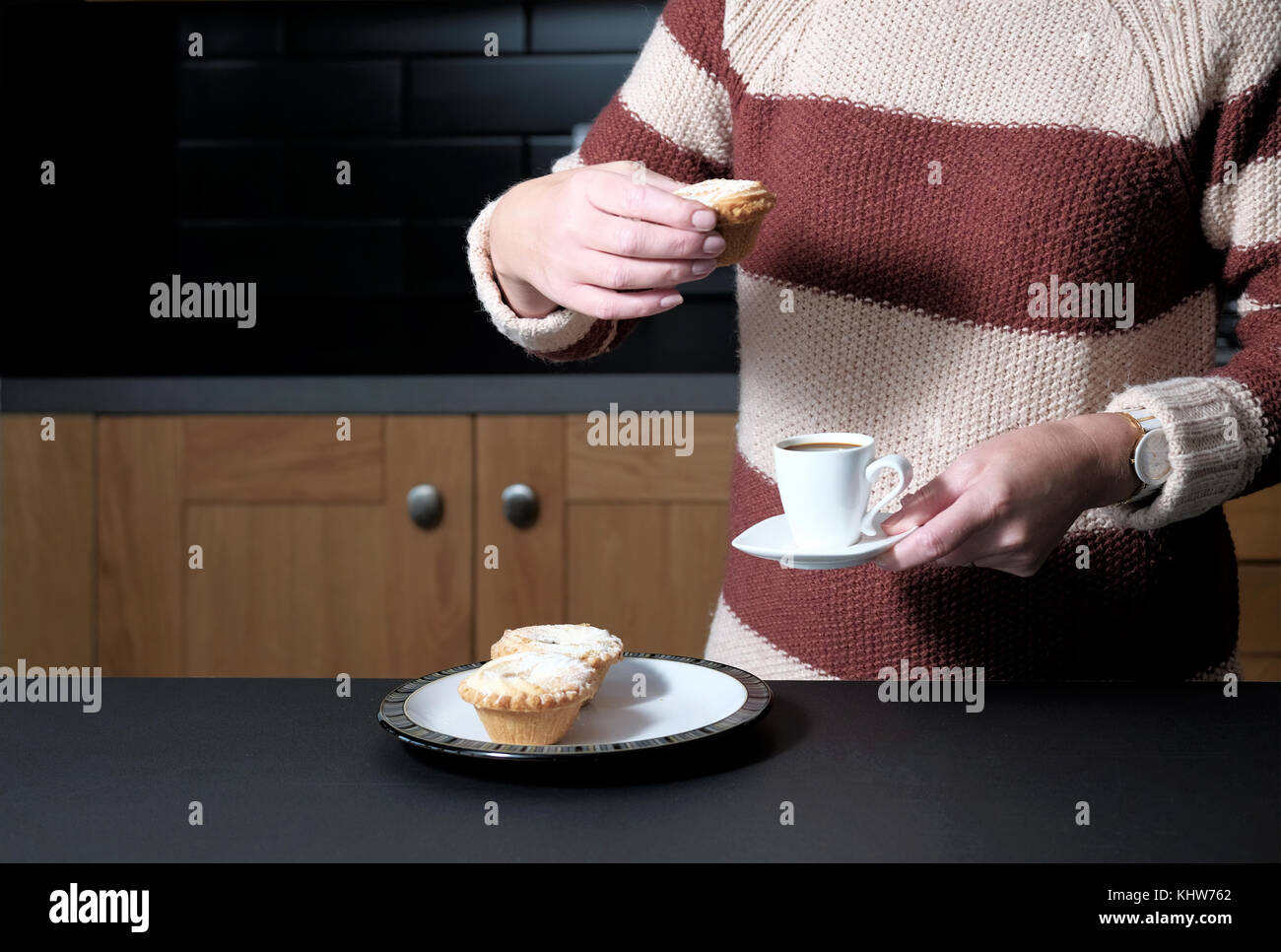 A smart but casually dressed woman holding an espresso coffee and a mince pie whilst standing in her kitchen at home Stock Photo