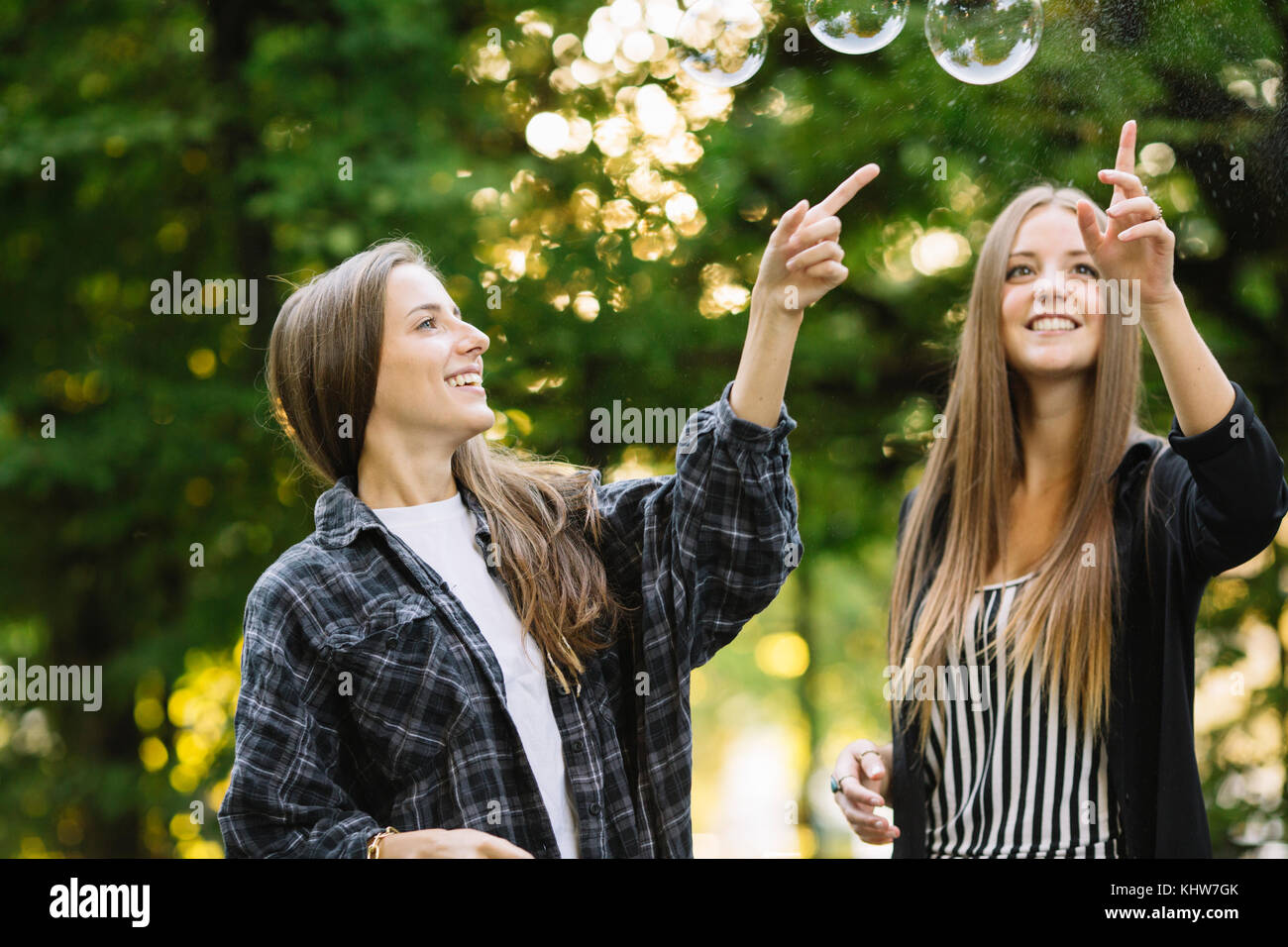 Two young female friends bursting floating bubbles in park Stock Photo