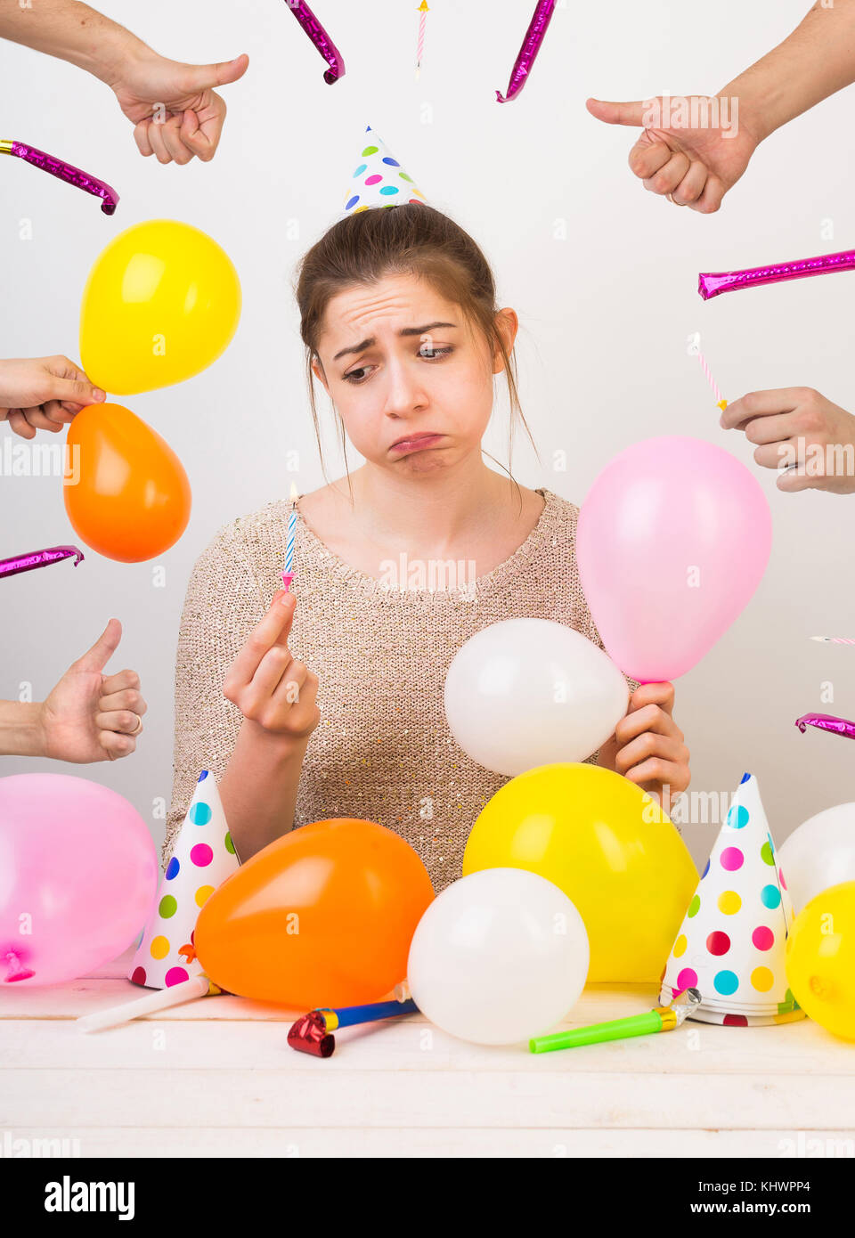 emotions, sentiments, birthday party concept. very sad disheveled girl, that is sulking, she is surrounded by colorful balloons, party hats with dots, hands with thumbs up and whistles Stock Photo
