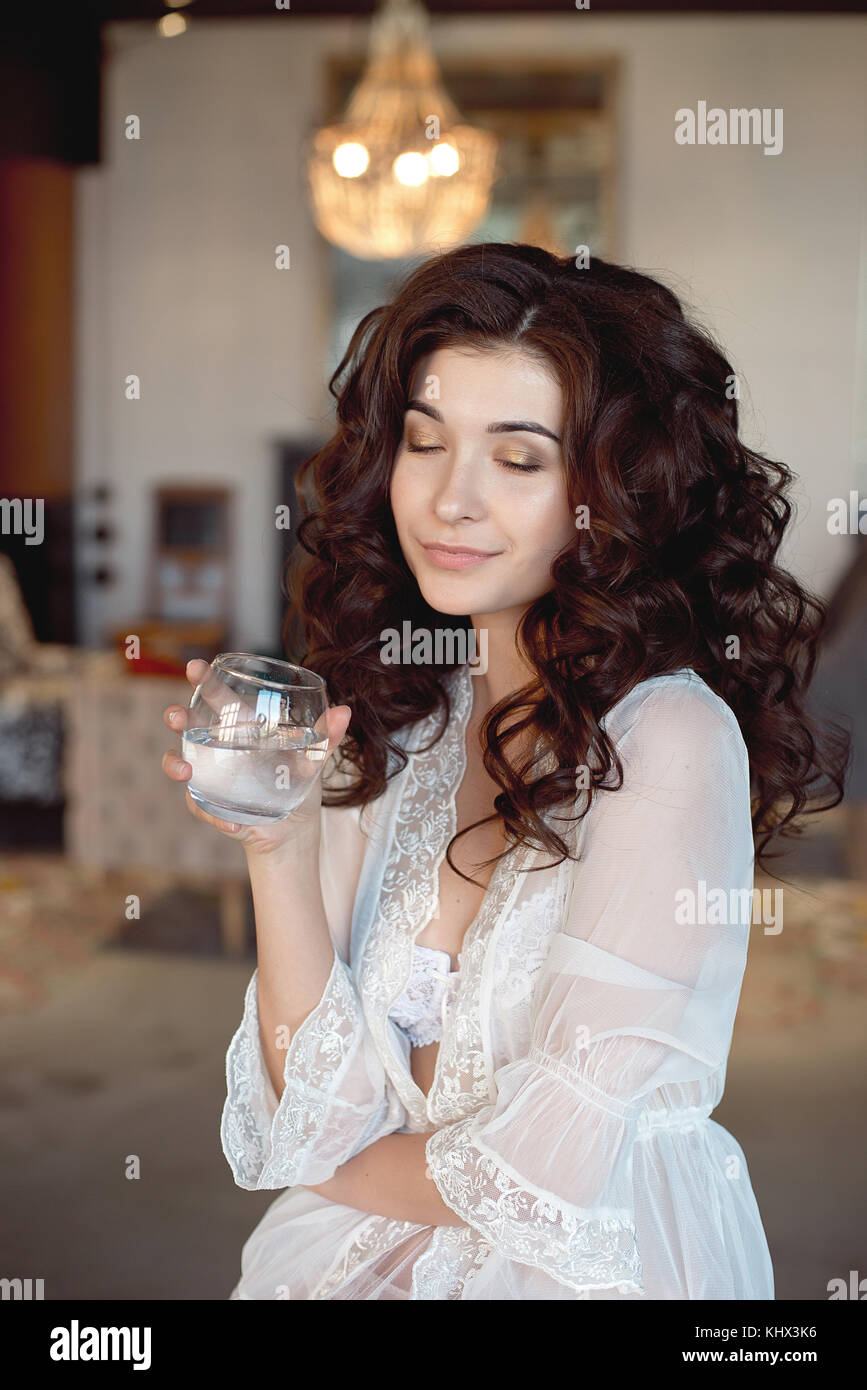 Attractive coy woman with downcast eyes wearing eye shadow holding a glass of cold beverage in her hand as she sits smiling on a sofa at home with a s Stock Photo