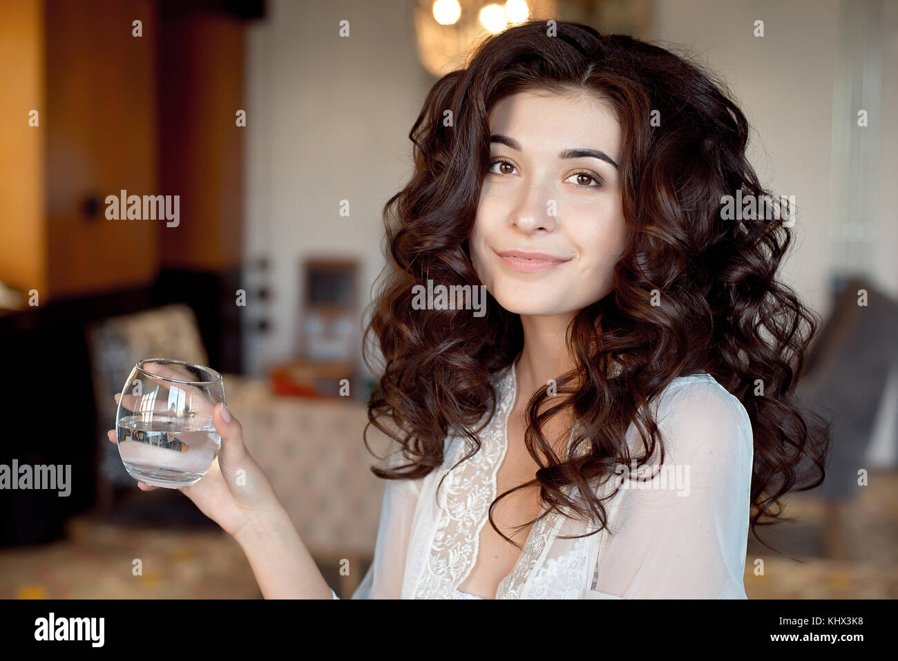 Attractive coy woman with downcast eyes wearing eye shadow holding a glass of cold beverage in her hand as she sits smiling on a sofa at home with a s Stock Photo