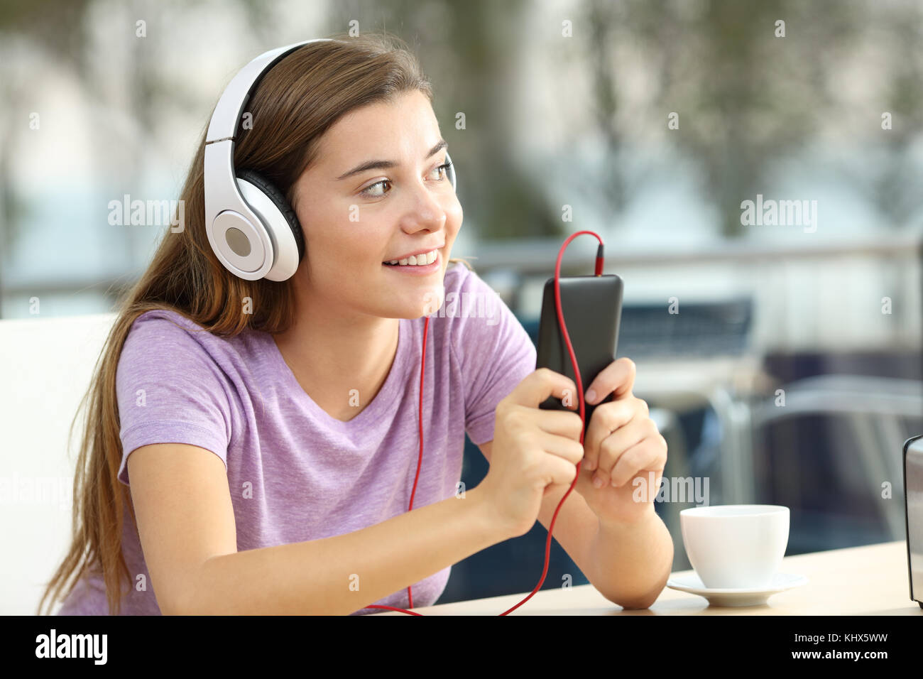 Happy teen listening to music from a smart phone wearing headphones and looking at side in a coffee shop Stock Photo