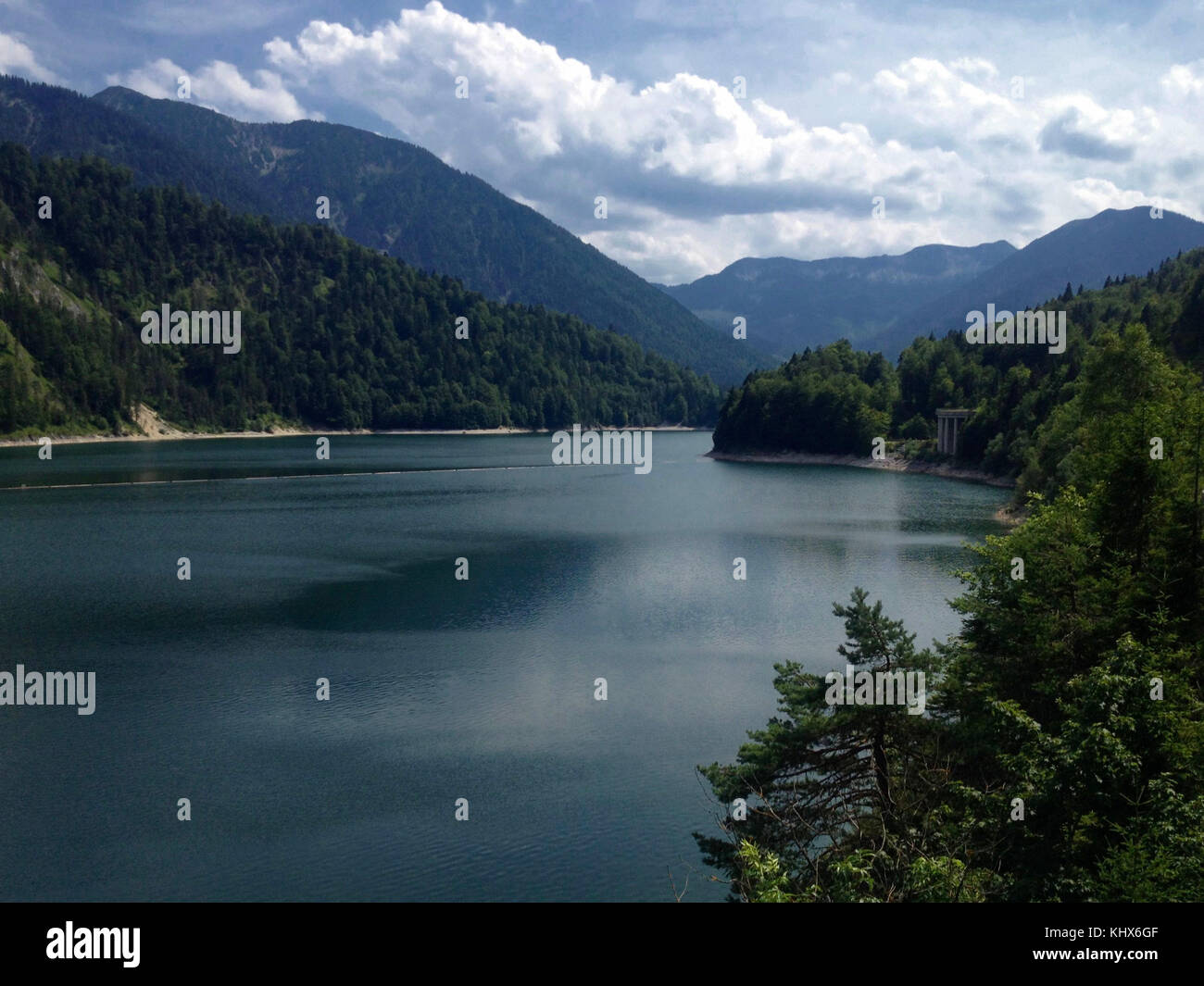 Peaceful freshwater lake at the Bavarian alps. Framed with the typical mountains and trees at a sunny day with blue sky and clouds. Stock Photo