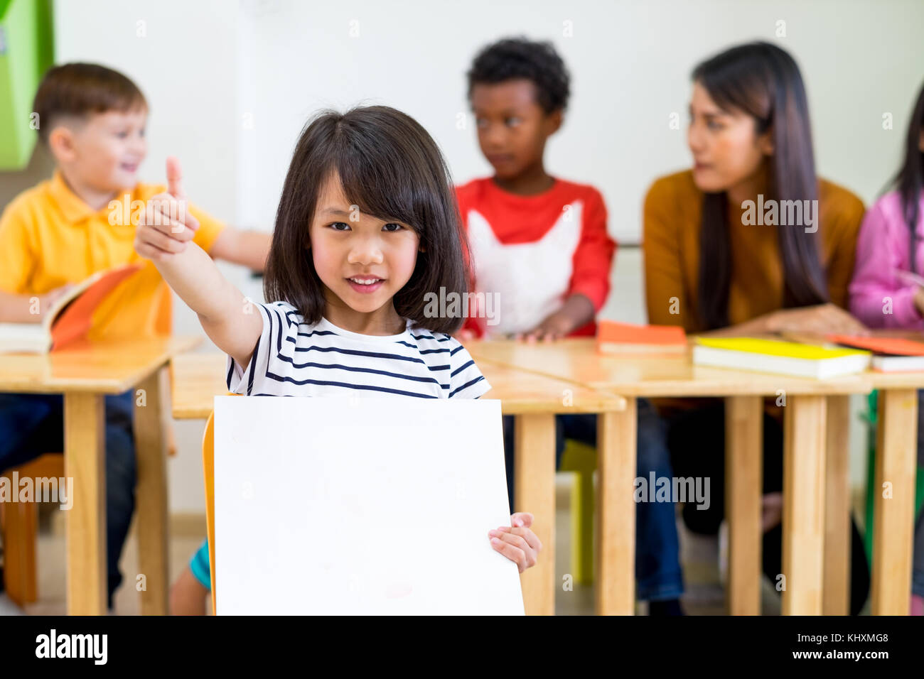 Kid girl thumbs up and holding blank white poster with diversity friends and teacher at background,Kindergarten preschool,mock up for adding text.chil Stock Photo