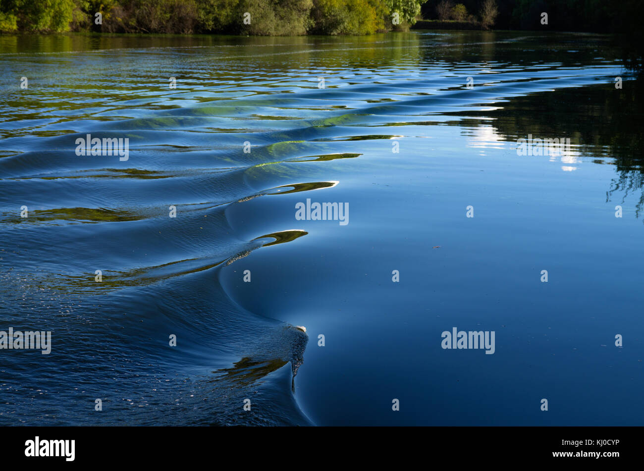 Boat wake curving in a river of calm water reflecting riverbanks and blue sky. Stock Photo