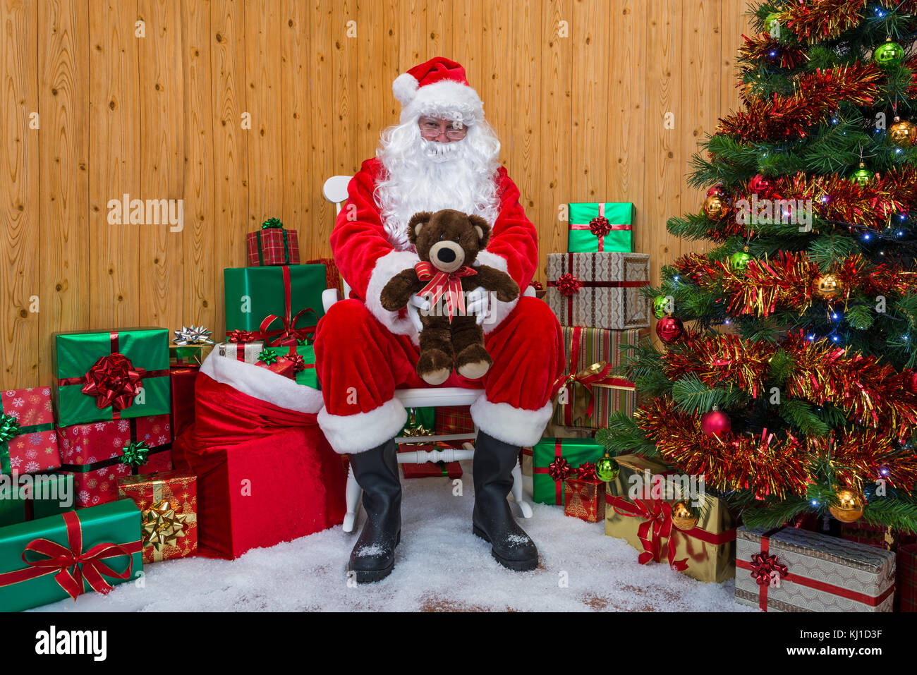 Santa Claus in his grotto surrounded by a Christmas tree with presents and gift wrapped boxes offering you a teddy bear. Stock Photo