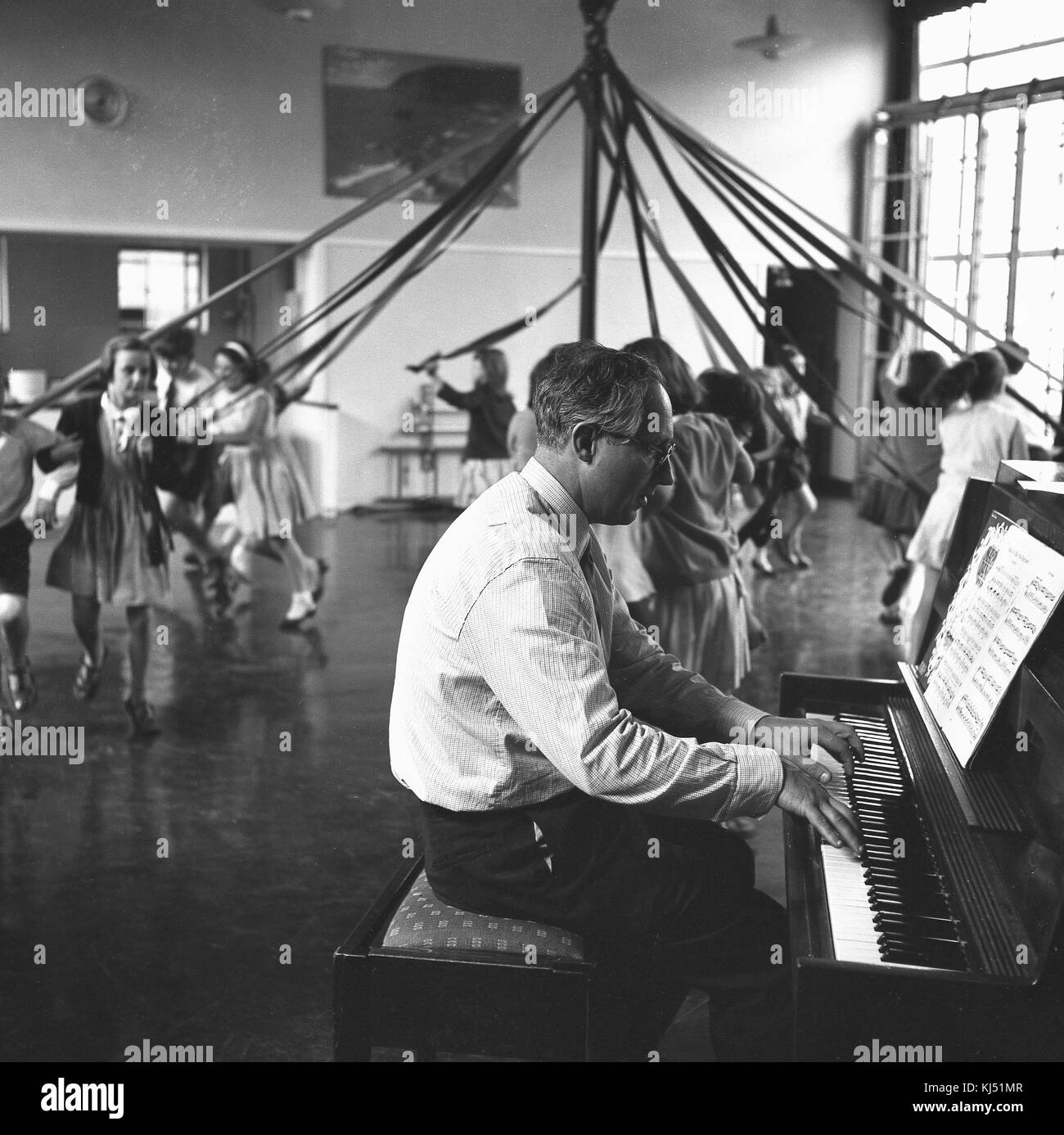 1950s, historical, inside a school hall, a male teacher plays the piano as primary school children skip and dance around a maypole holding ribbons, England, UK. Stock Photo