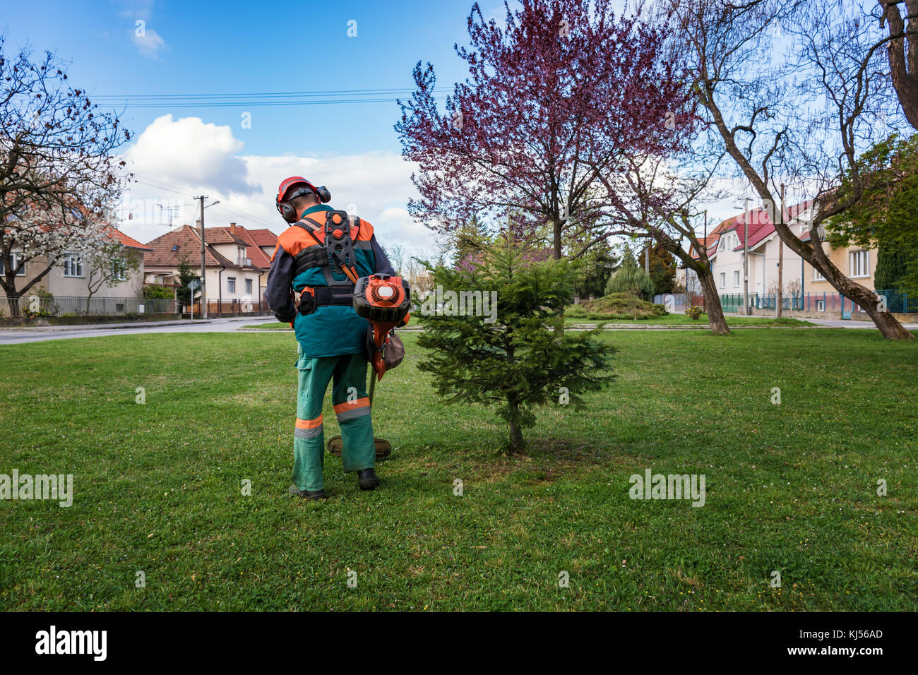 Man in overall and safety helmet trims overgrown grass by grass cutter Stock Photo